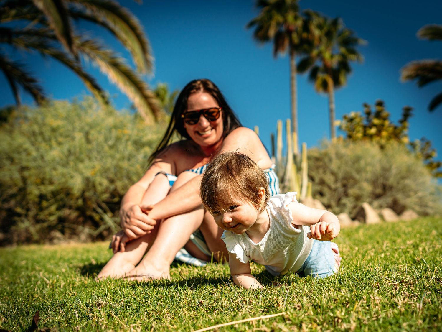 Smiling mother with sunglasses sitting on grass with toddler near tropical palm trees.