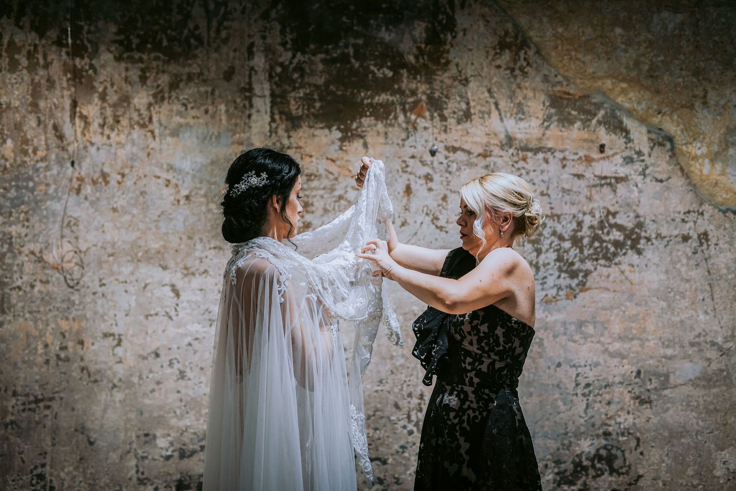 Bride in lace wedding gown with cape being assisted by woman in black dress against rustic wall.
