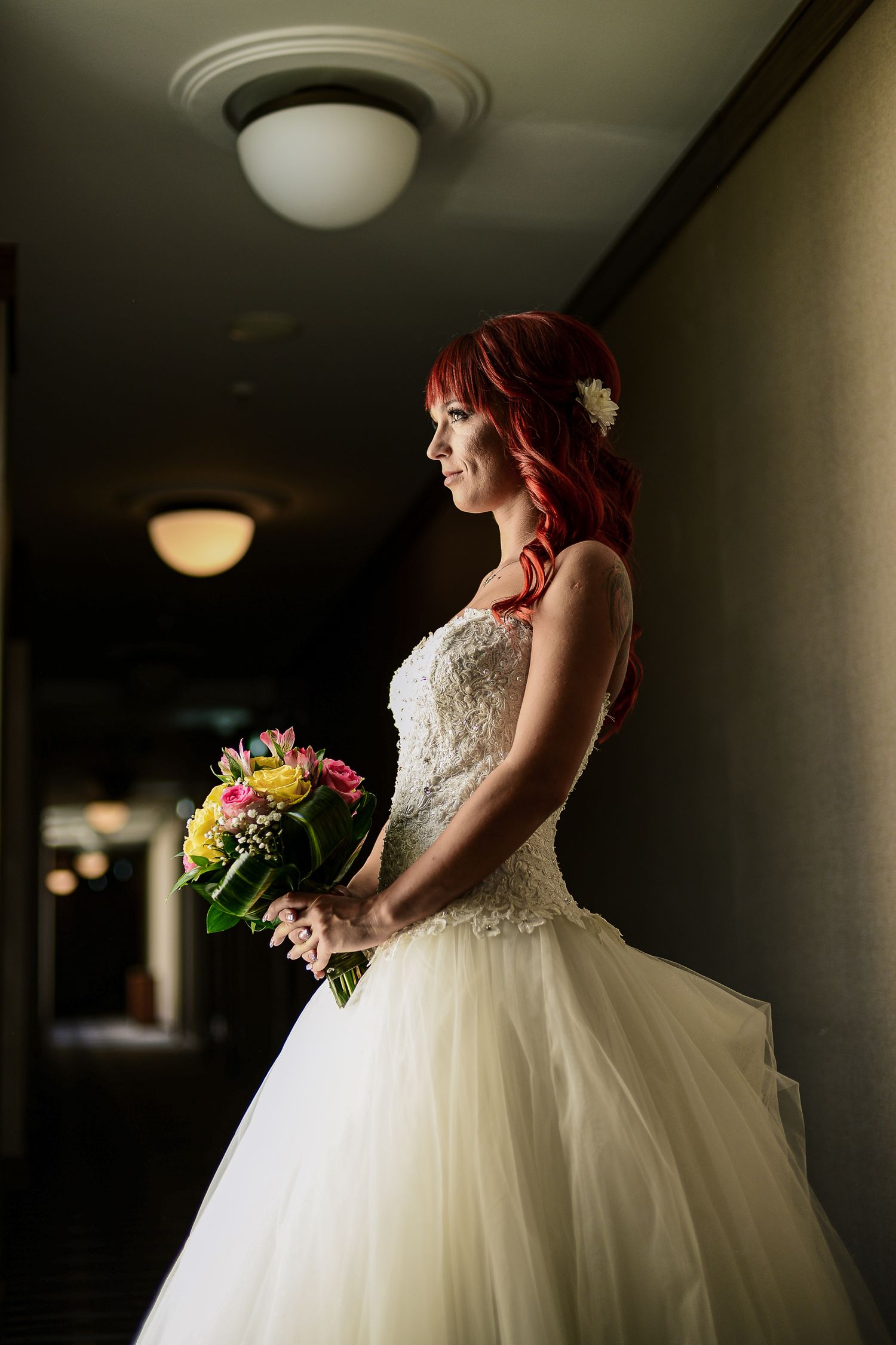 Stunning red-haired bride in lace ballgown holds colorful bouquet while gazing through a hotel corridor.