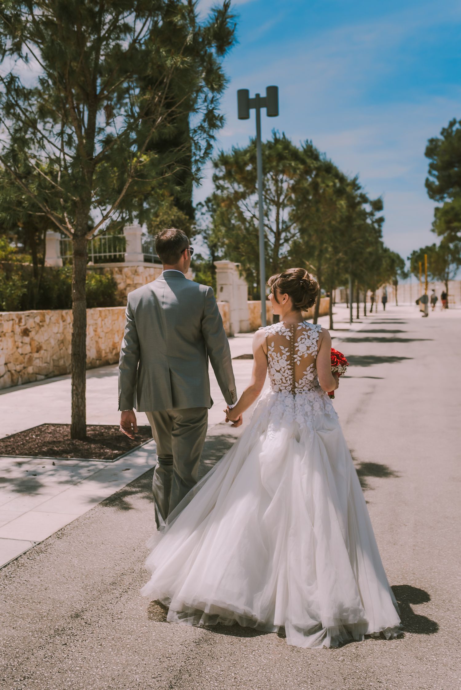 Bride and groom walking hand in hand along a sunny tree-lined path after their wedding ceremony.