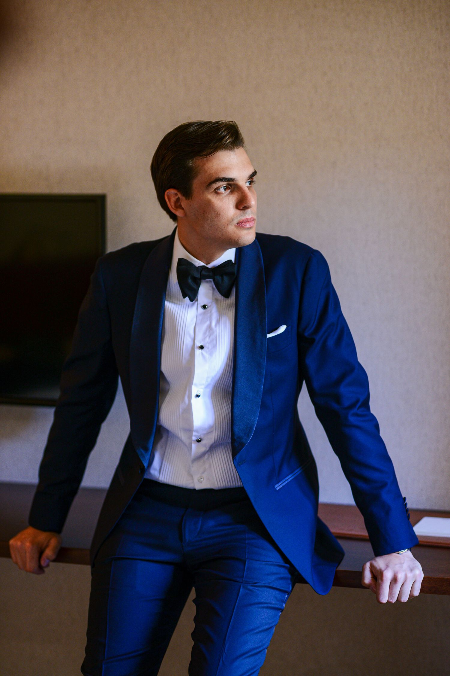 Elegant groom in a blue tuxedo with black bow tie leans against a railing in a sophisticated indoor setting.