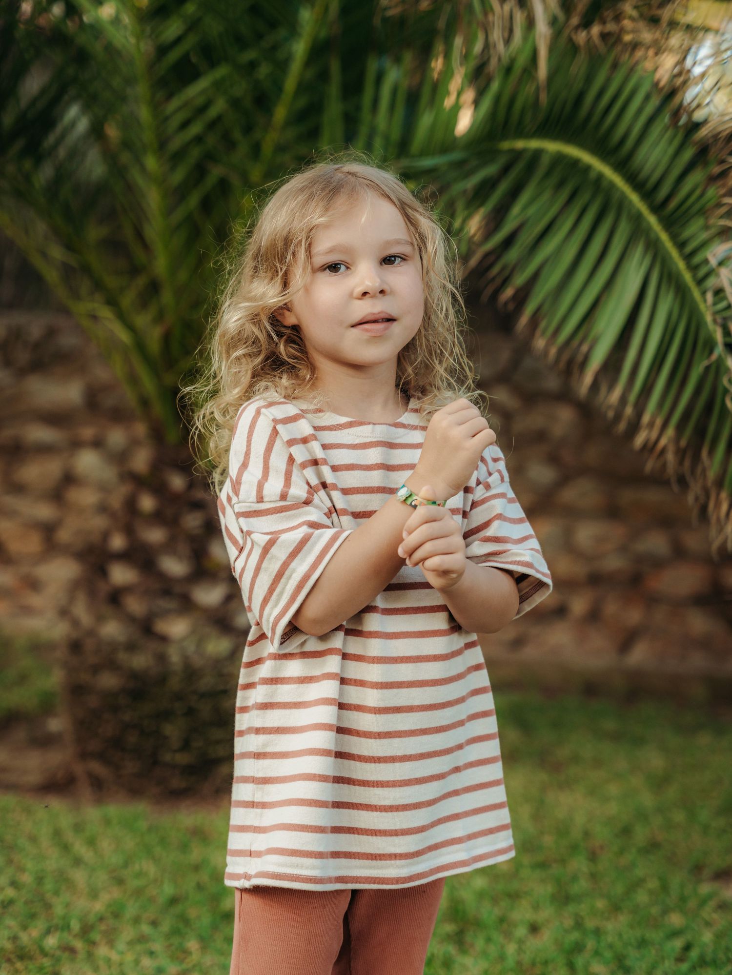 Cute blonde toddler girl in striped dress standing near palm tree in garden.