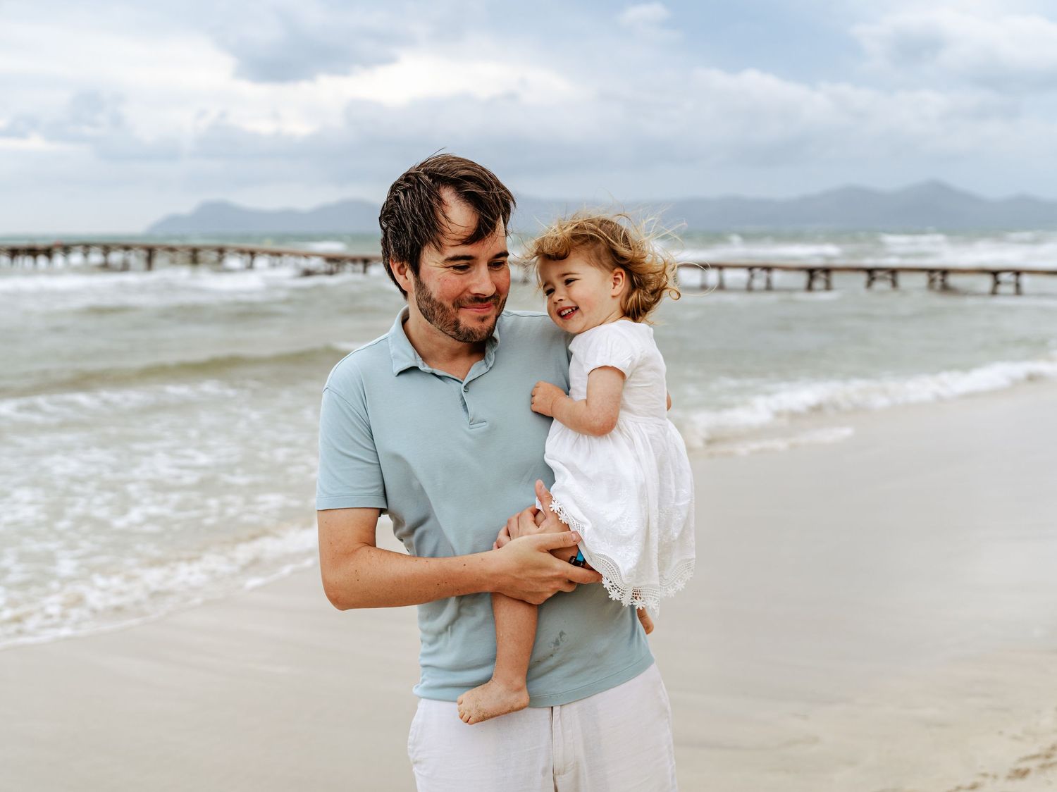 Smiling dad in blue polo holding happy toddler by sea.