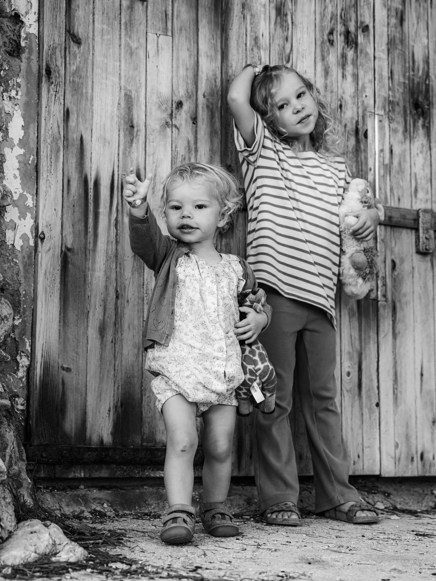 Black and white photo of two young children posing by a rustic wooden door with a stuffed giraffe.