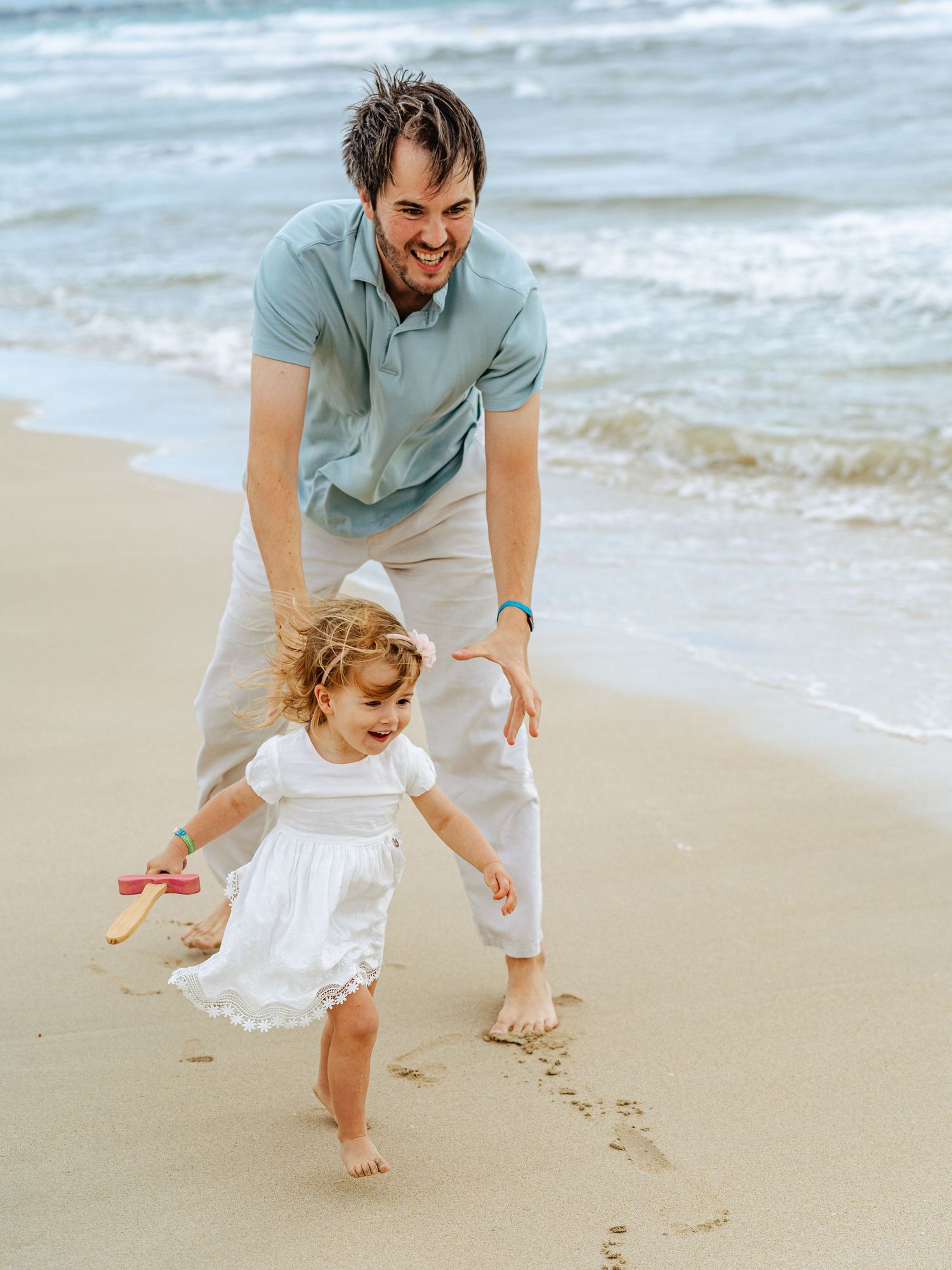 Father chasing his happy toddler daughter running barefoot on a sandy beach shoreline.