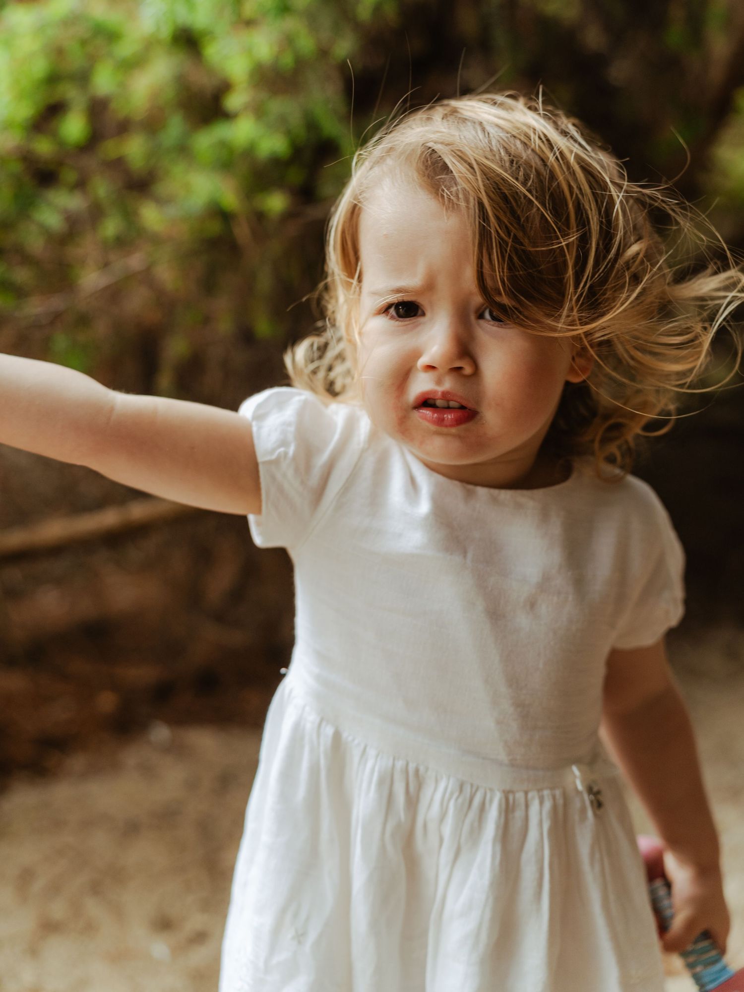 Toddler girl in white dress with windblown hair reaching out in outdoor natural setting.