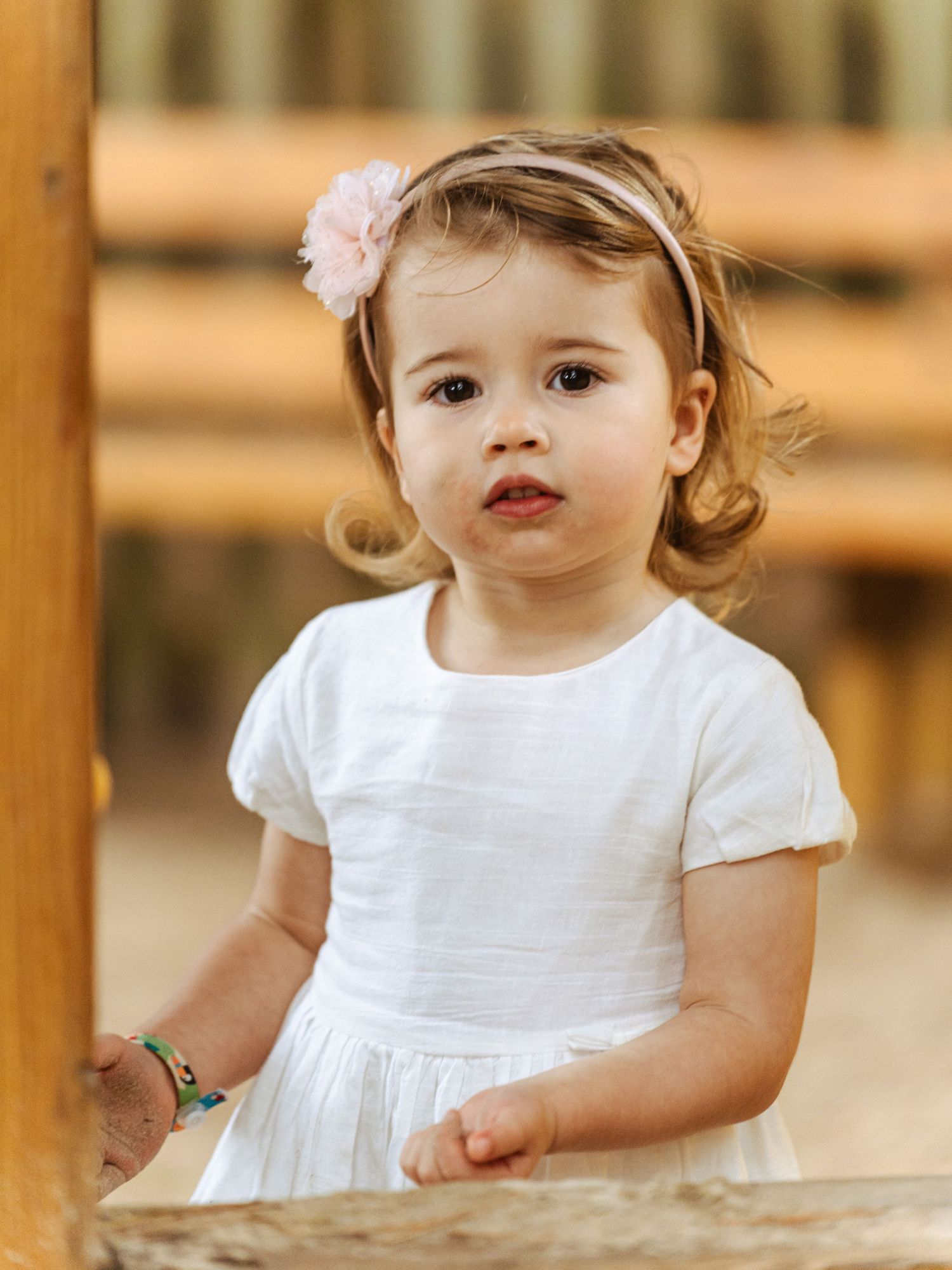 Adorable toddler girl in white dress with pink flower headband standing at wooden playground.