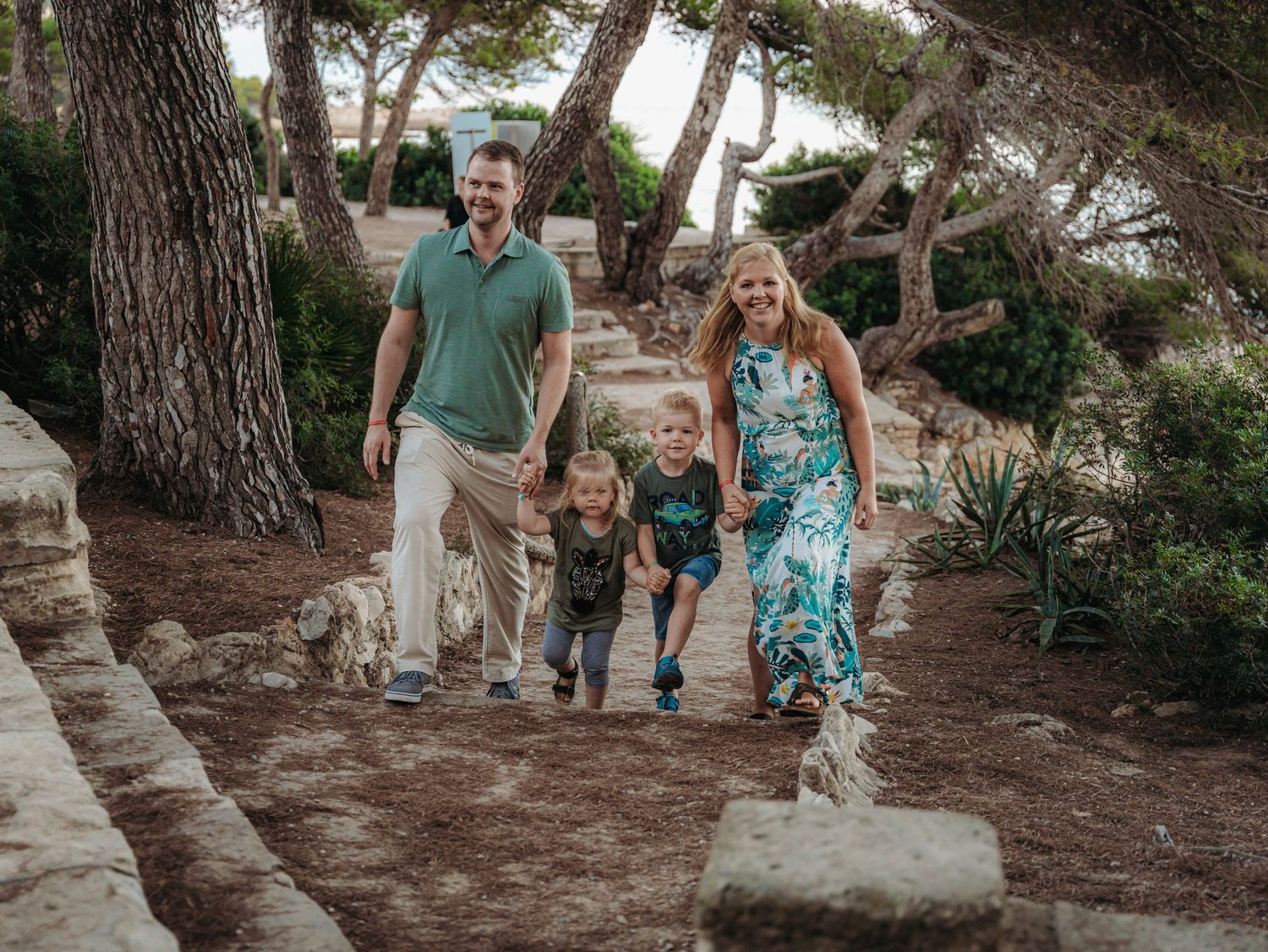 Happy family of four walking together on scenic stone path surrounded by Mediterranean pine trees.