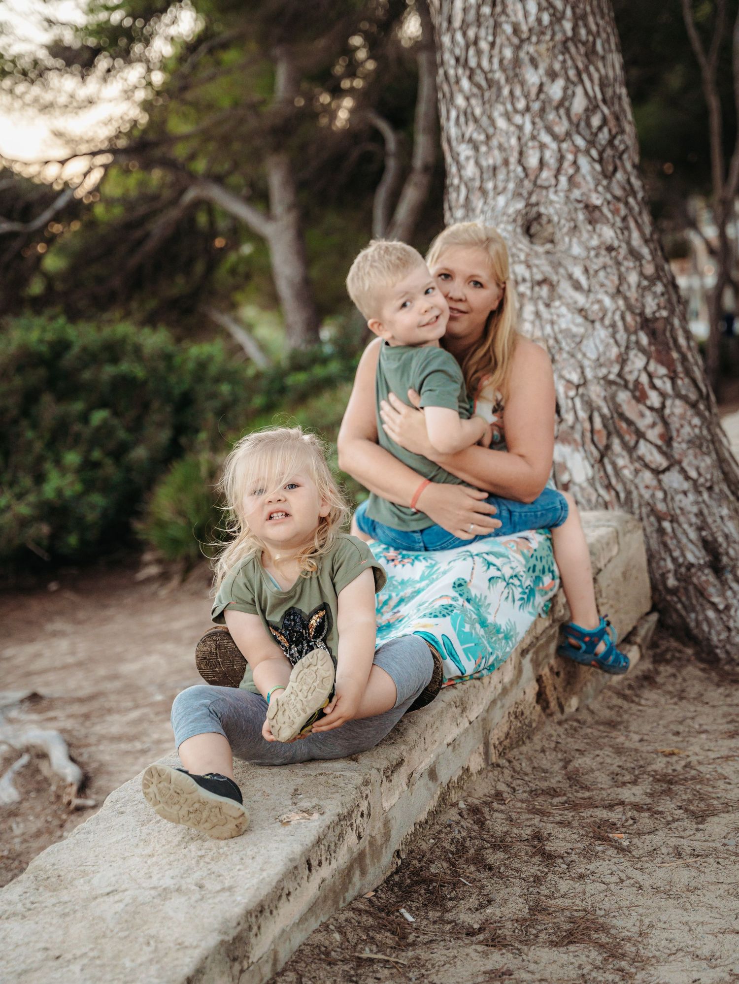 Mother sitting under pine tree with two young blonde children on stone bench in wooded park.
