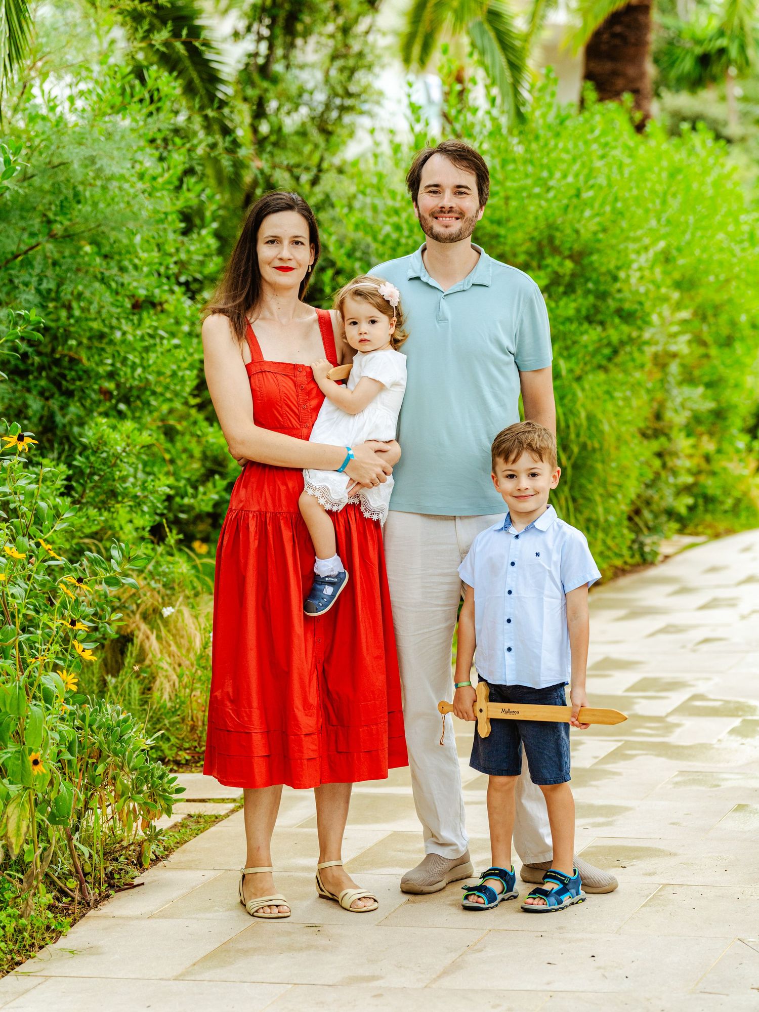 Happy family of four posing on garden path, mother in red dress.