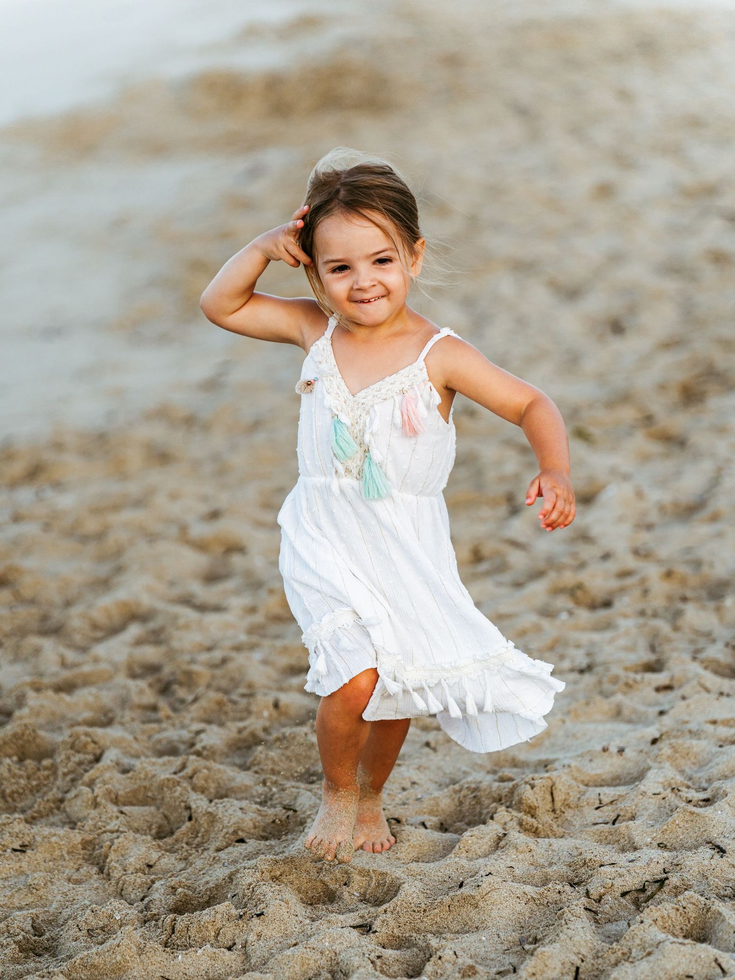 A cheerful young girl in a white boho dress dancing barefoot on a sandy beach.