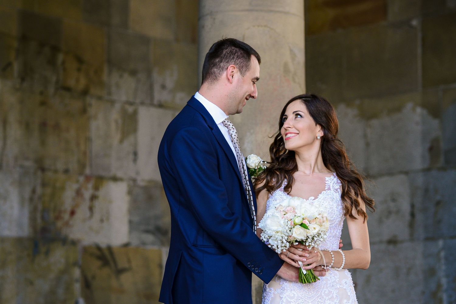 Elegant bride holding white bouquet smiling at groom in blue suit beside stone columns.