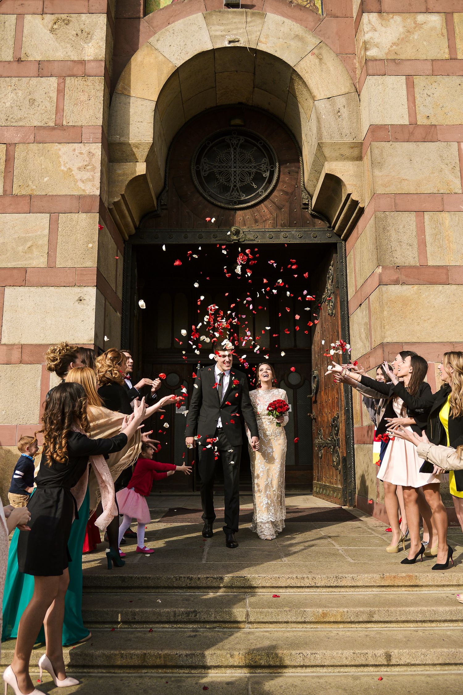 Newlyweds exit church doors as guests toss red and white flower petals in joyful wedding celebration.