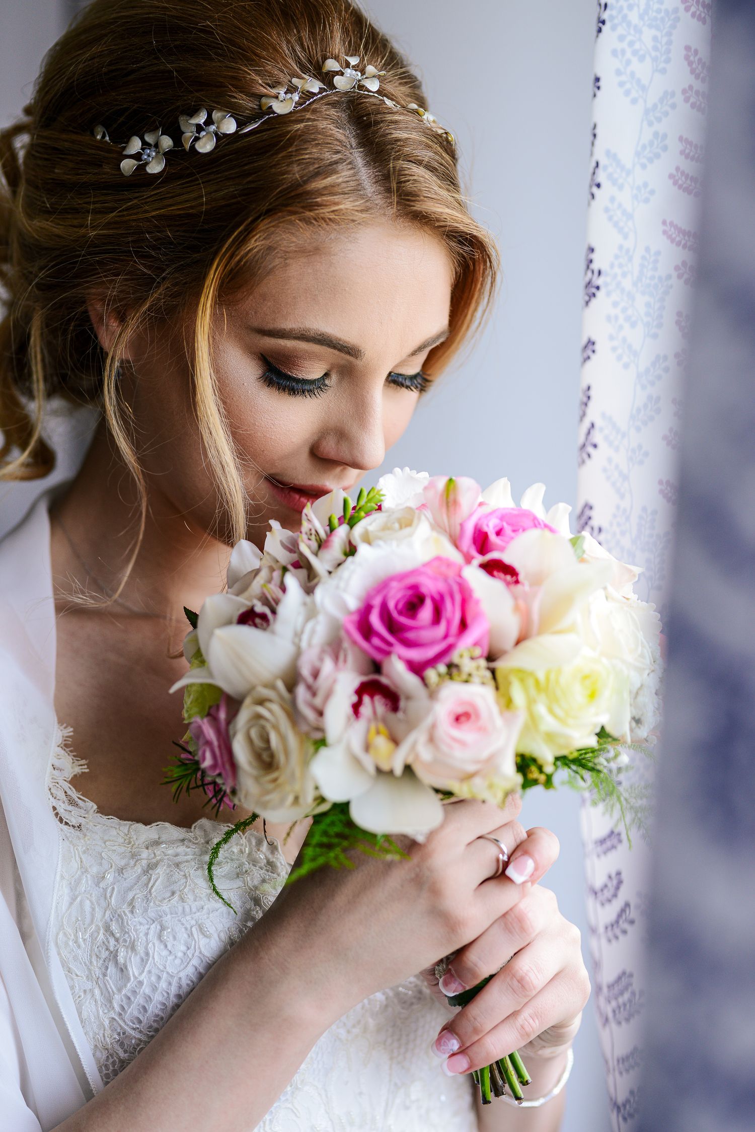 Elegant bride with floral hair accessory smells a colorful bouquet of pink and white roses by a window.