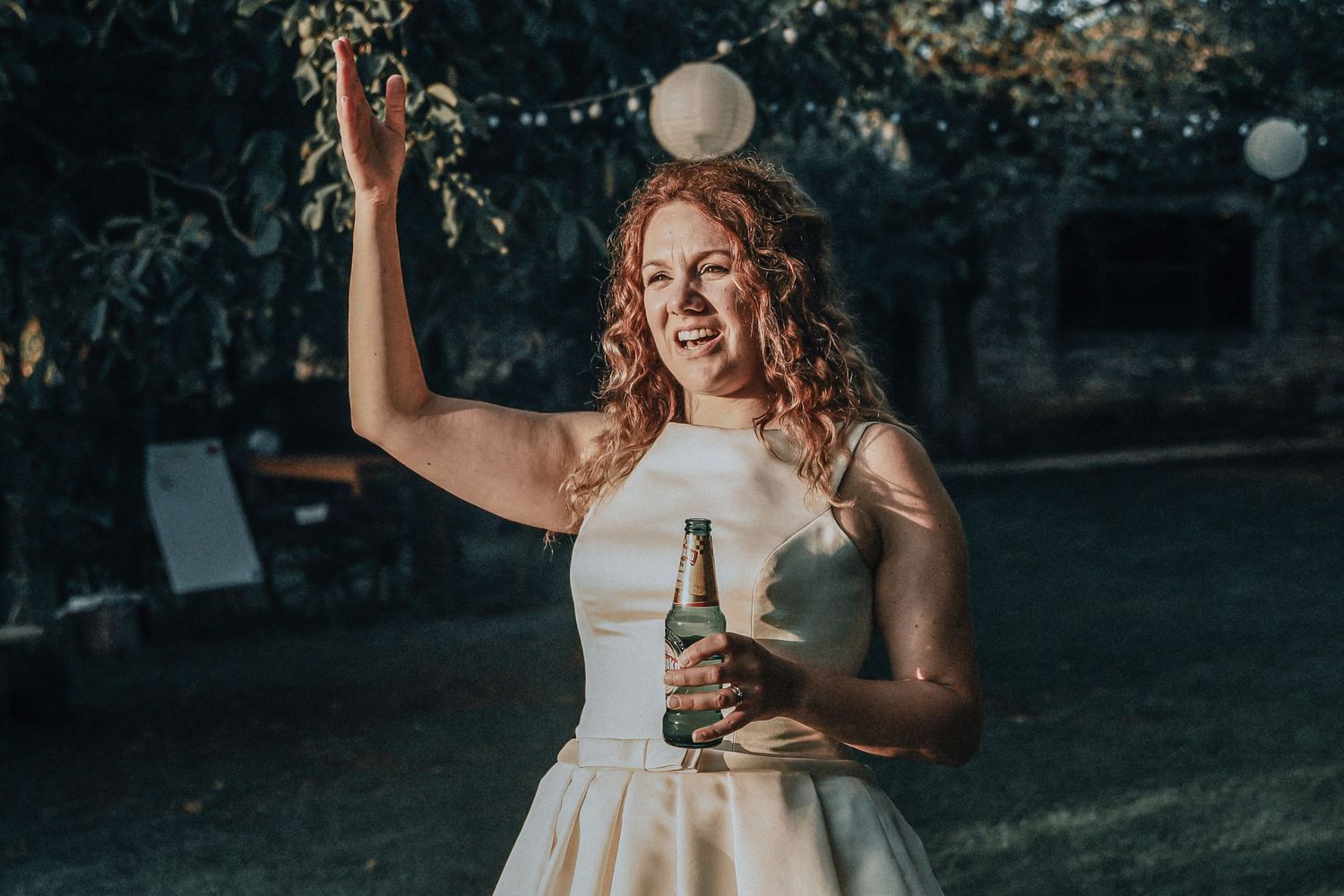 Smiling bride in white dress tossing a ball while holding a beer bottle at an outdoor wedding reception.