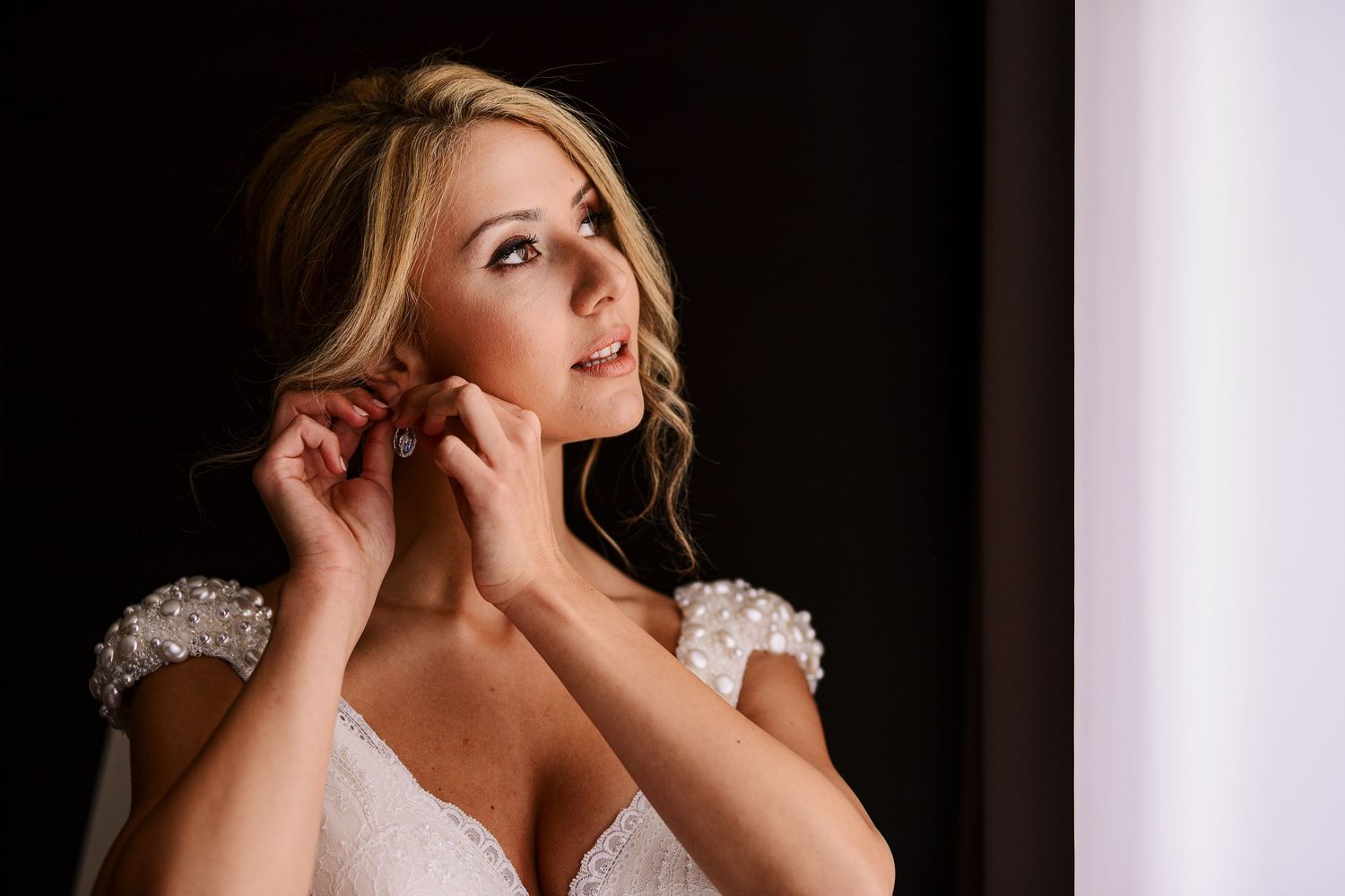 Elegant blonde bride in a lace wedding dress adjusting her earring near a bright window on her wedding day.