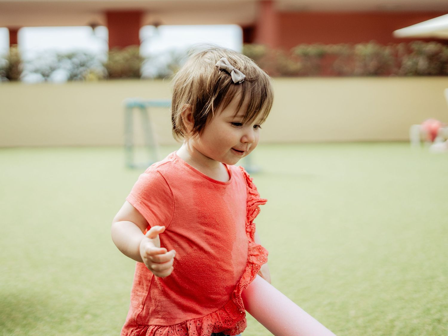 Adorable toddler girl in red top with hair clip playing outdoors on green grass.