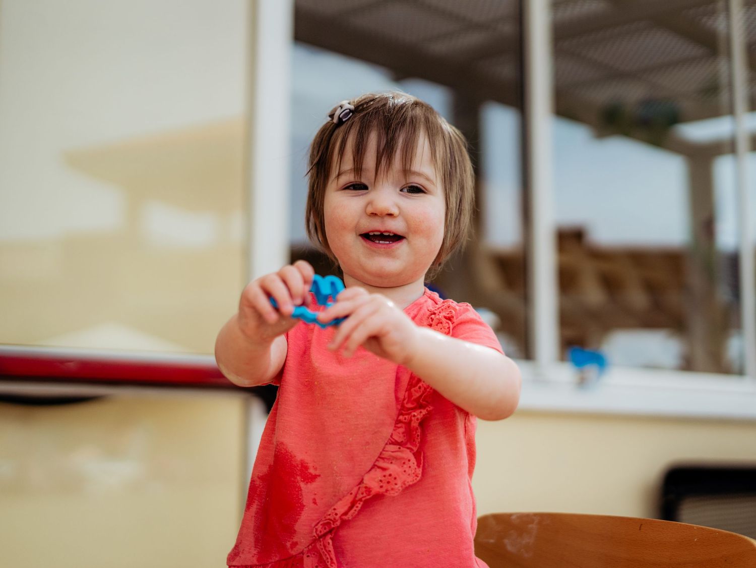 Happy toddler girl in coral shirt smiling and holding a blue toy outdoors near a yellow wall.