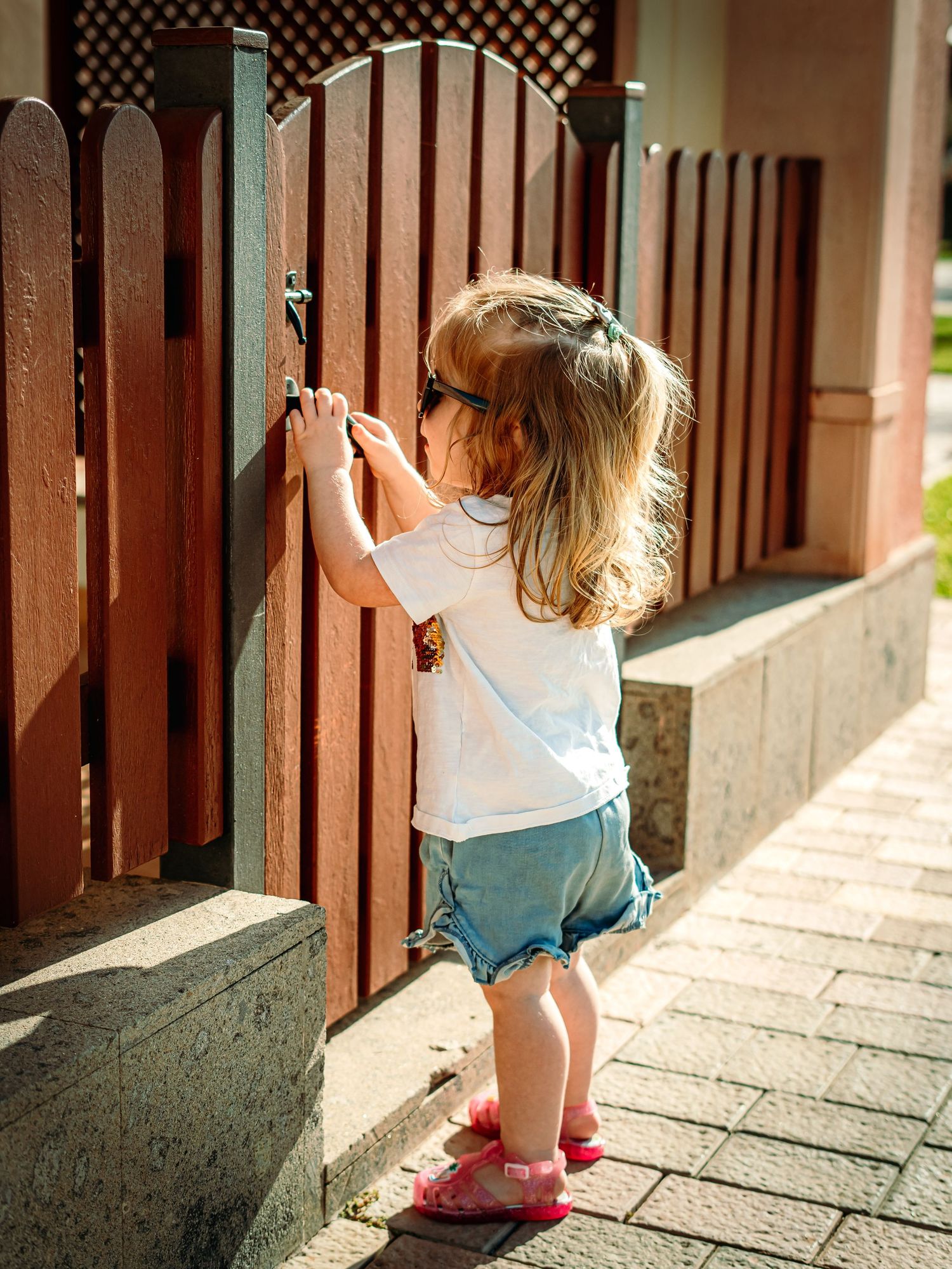 Young toddler girl in white tee and denim shorts with sunglasses opening a brown wooden garden gate.