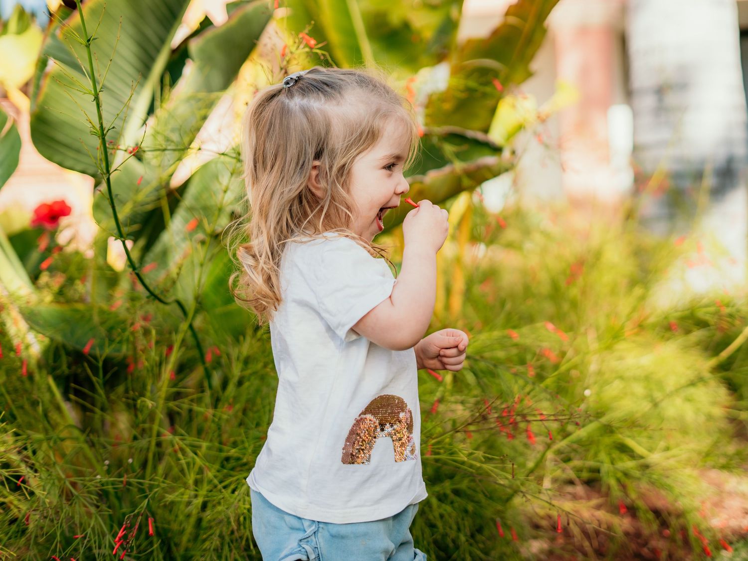 Cute blonde toddler girl in a rainbow t-shirt exploring lush tropical garden greenery.