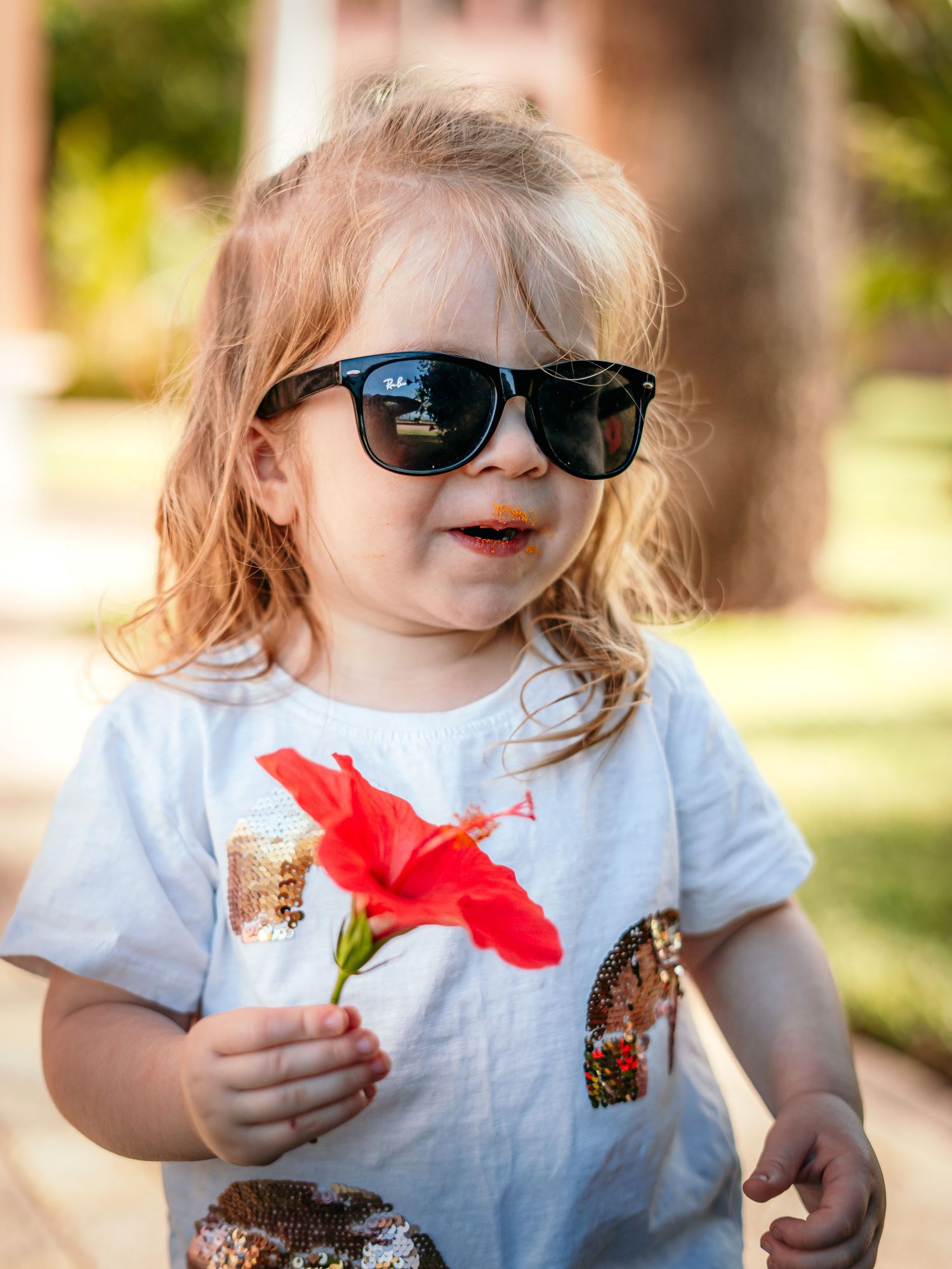Cute toddler wearing black sunglasses holds a red hibiscus flower while standing outdoors.