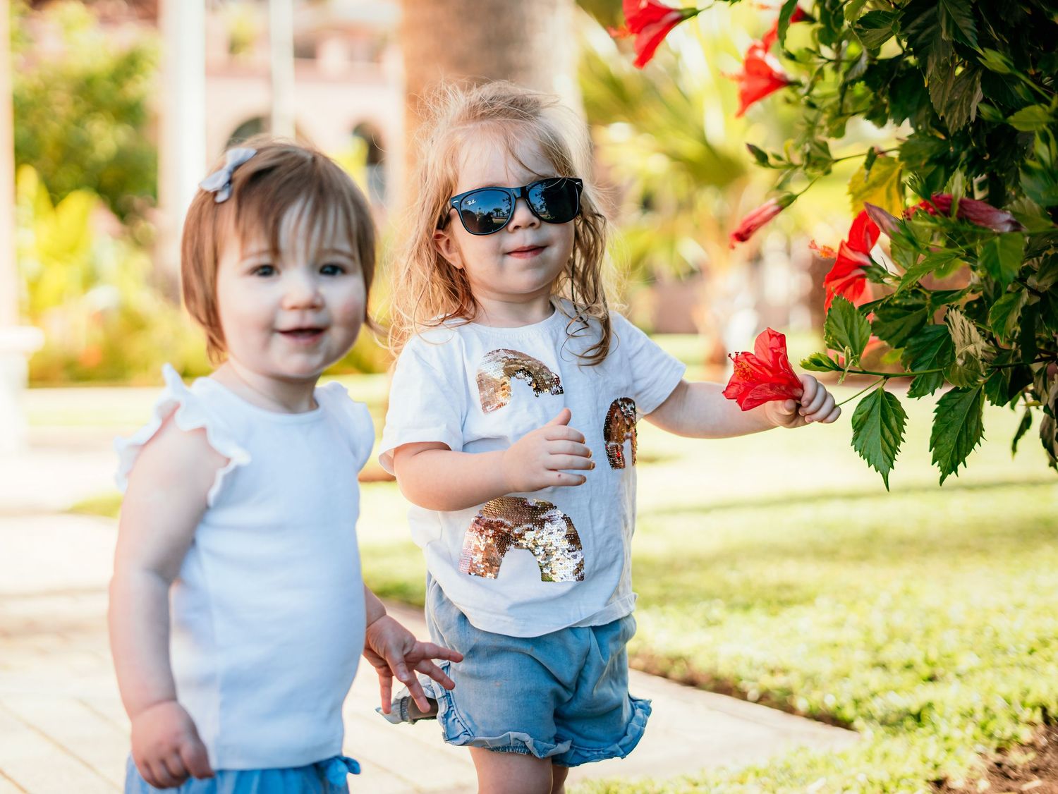 Two young sisters stand together near red hibiscus flowers, one wearing sunglasses in sunny park.