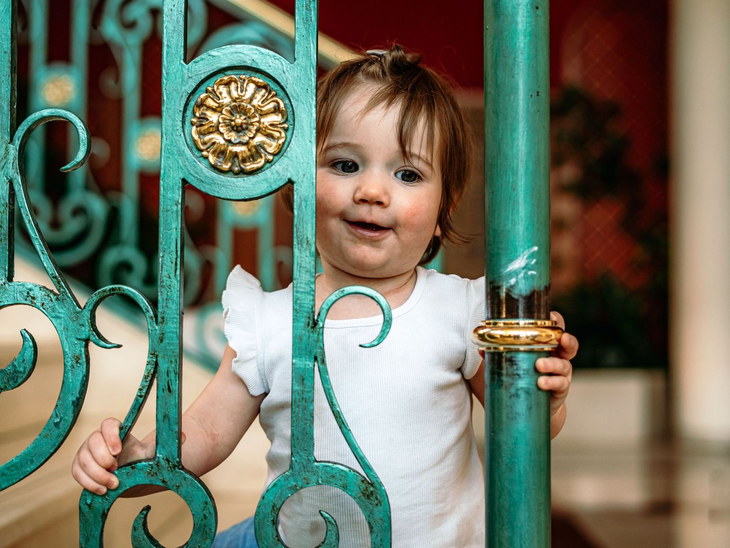 A cheerful baby girl in a white dress peeks through decorative green and gold iron railings.
