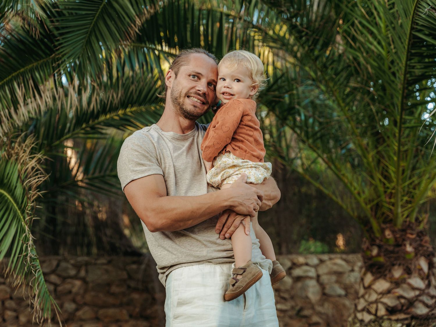 Happy father smiling while holding young toddler outdoors surrounded by tropical palm trees.