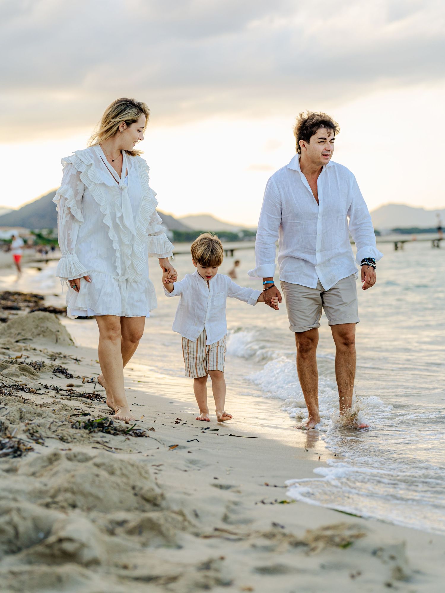 Parents and young son holding hands walking along a beach shoreline at golden hour in white attire.