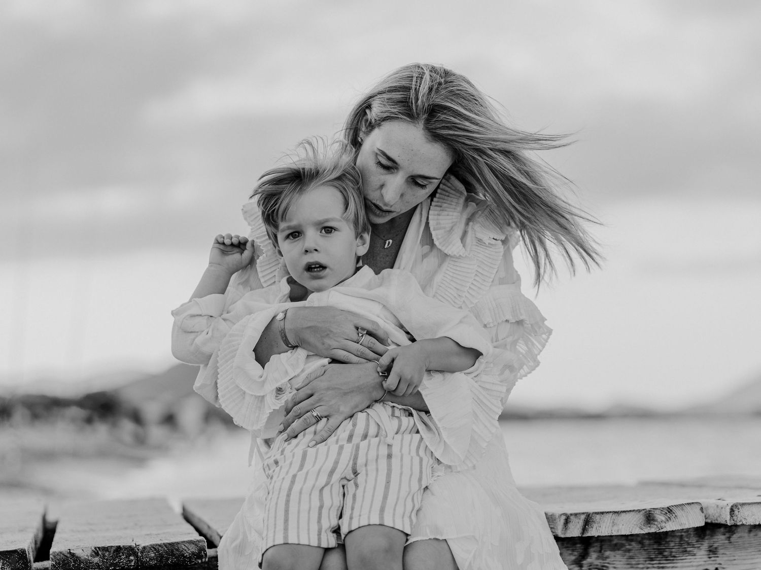 Black and white photo of mother embracing toddler on windy beach.