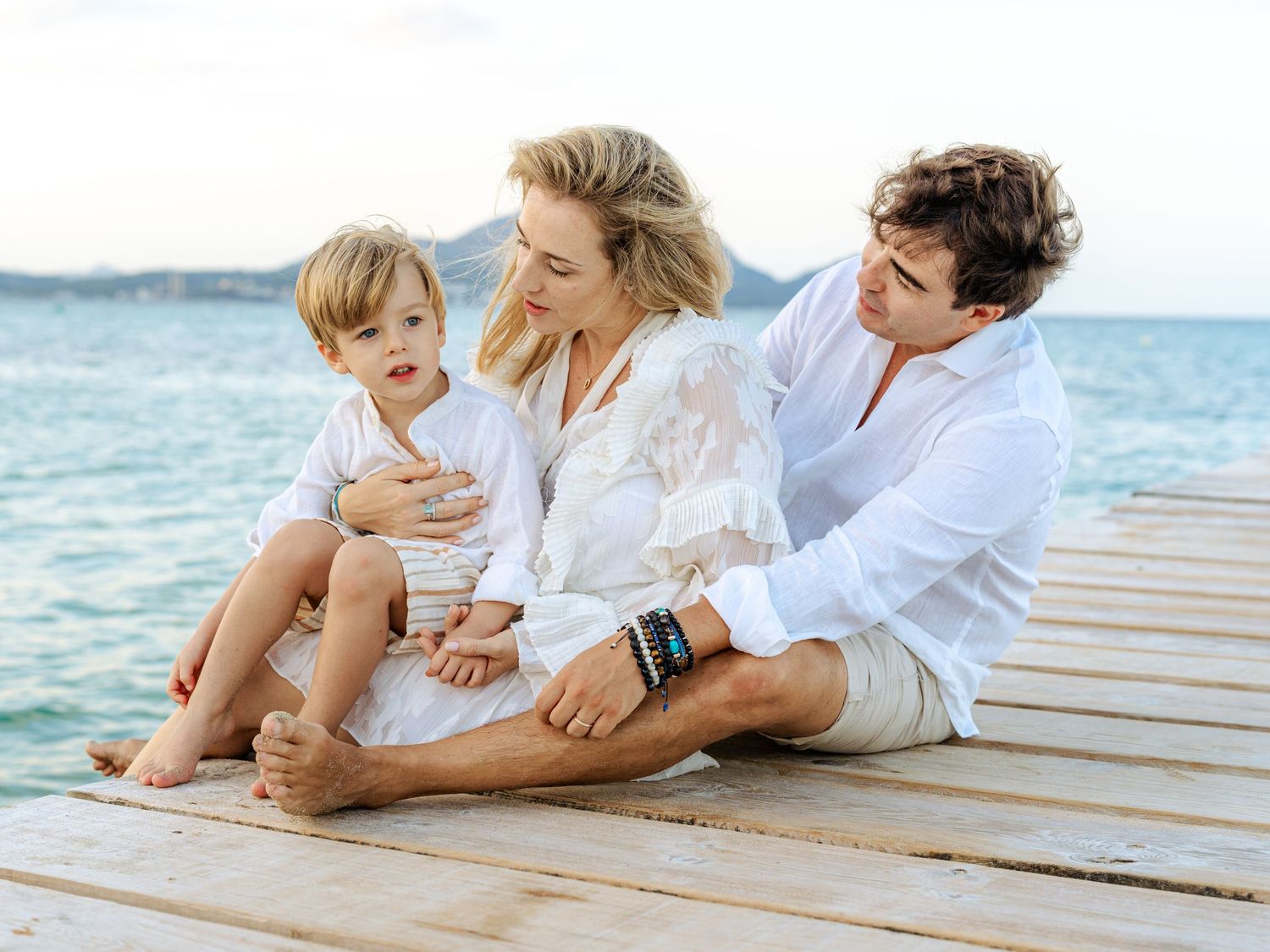 Loving family of three sharing a tender moment on a wooden pier with turquoise sea behind them.