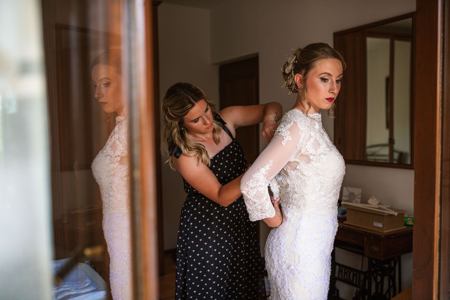 Bridesmaid zipping up bride's lace wedding dress reflected in mirror.