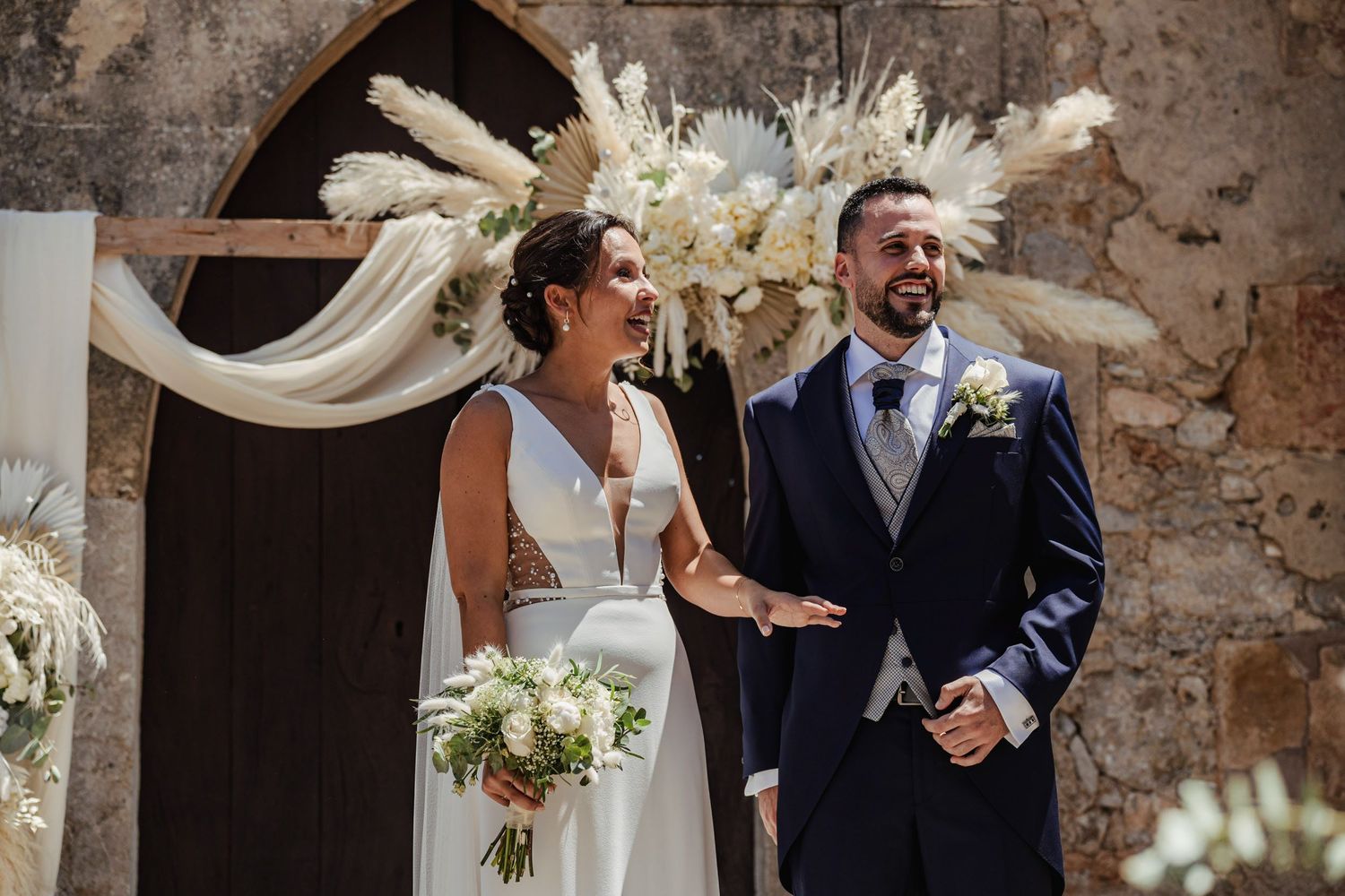 Bride and groom stand before boho floral arch with pampas grass inside rustic stone wedding venue.