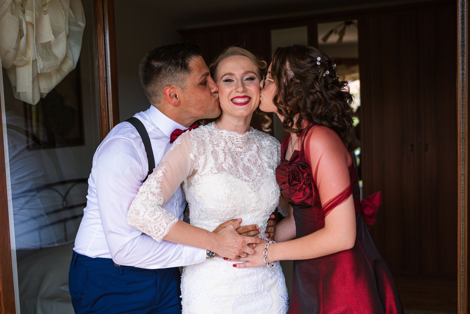 Happy bride in lace wedding dress receiving kisses on cheeks from man and woman in red dress.