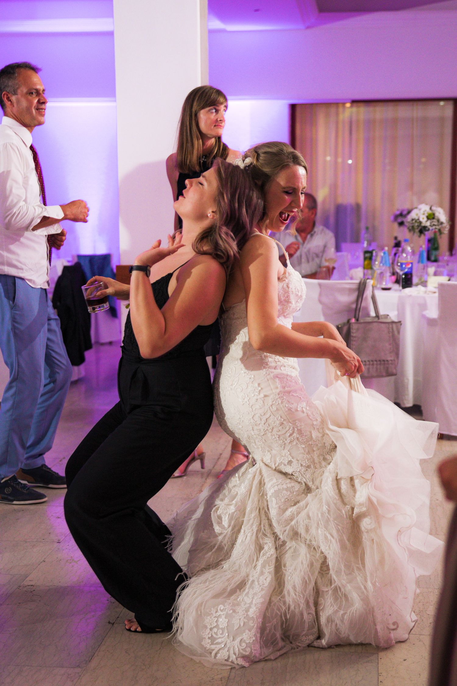 Bride in lace gown and guest in black dress dance together joyfully at wedding reception.