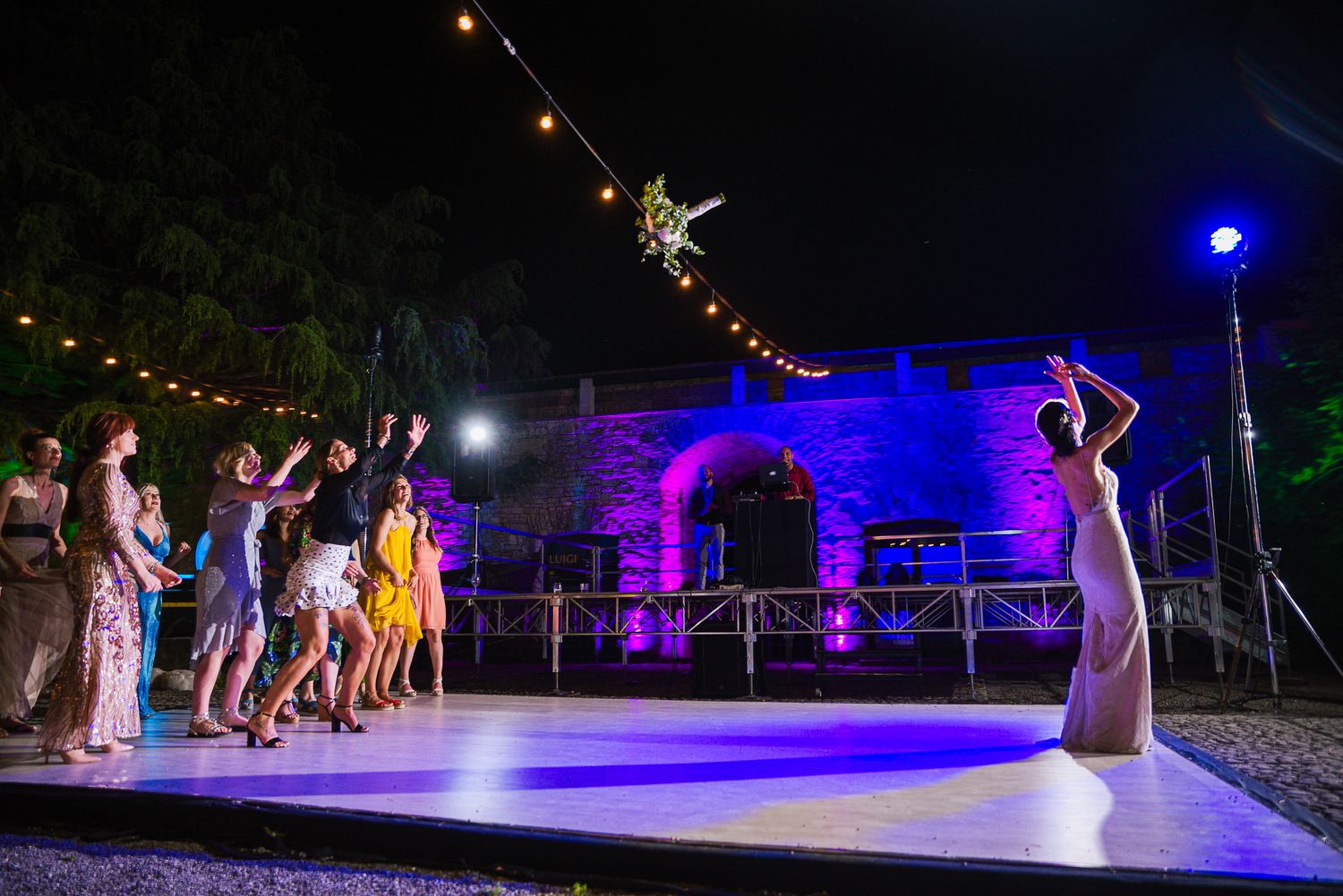 Bride tosses wedding bouquet to guests on an outdoor dance floor lit with colorful purple and blue lights.