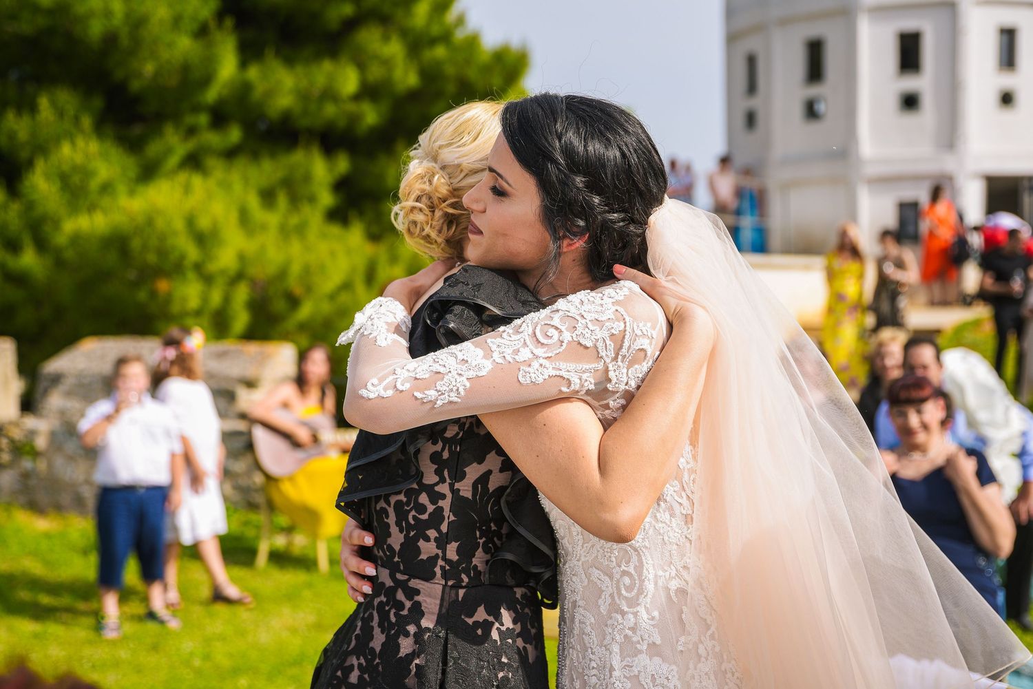 Bride in white lace gown embracing a guest in black lace dress at an outdoor wedding ceremony.