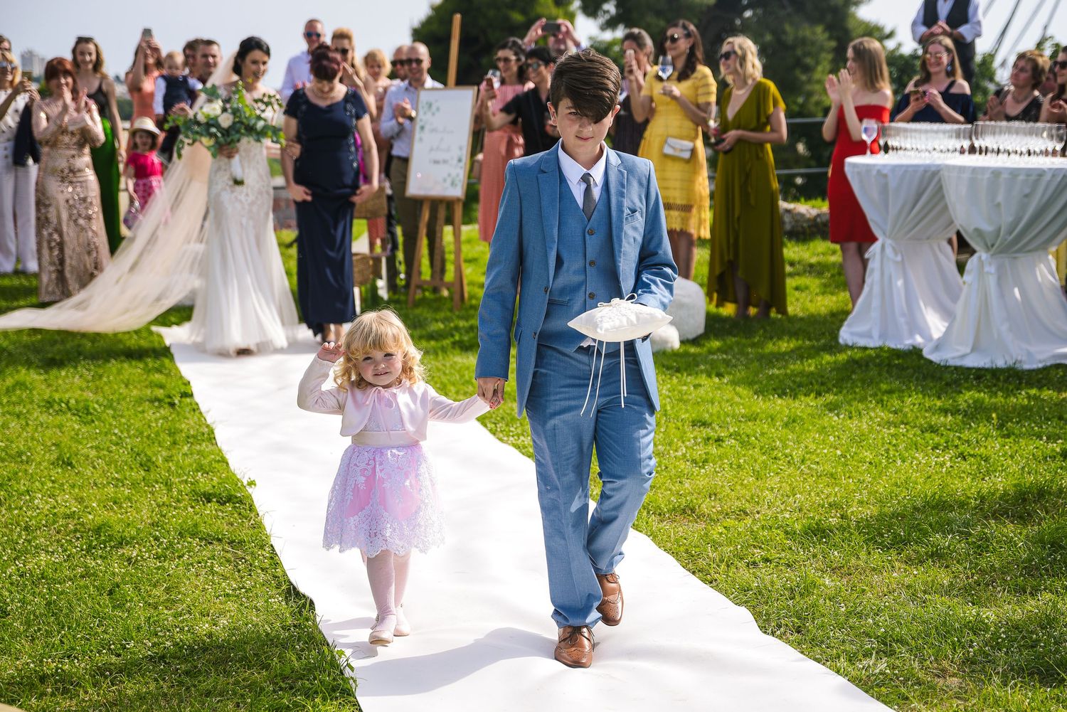 Young ring bearer in blue suit walks flower girl down white aisle at outdoor wedding.