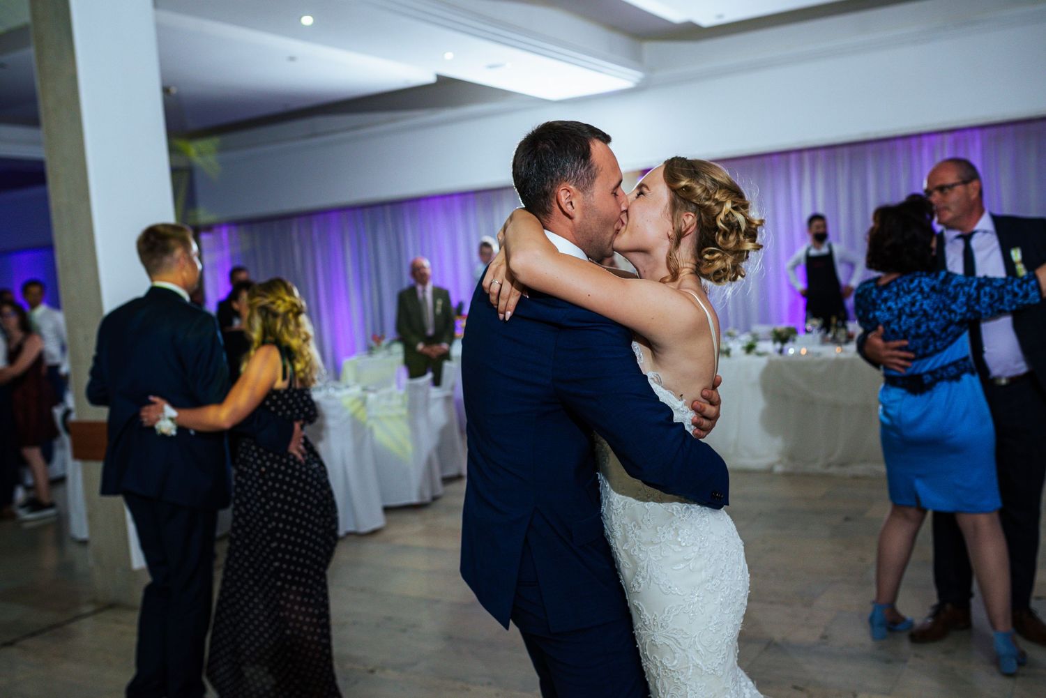 Bride and groom kissing while dancing together during their wedding reception surrounded by members of their family. Herzenshaus Studio.