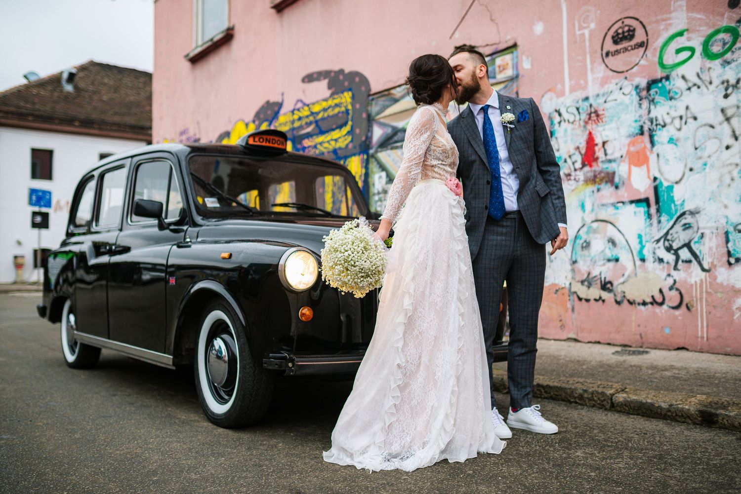 Bride and groom sharing a kiss beside a classic black London taxi cab with graffiti street art behind.