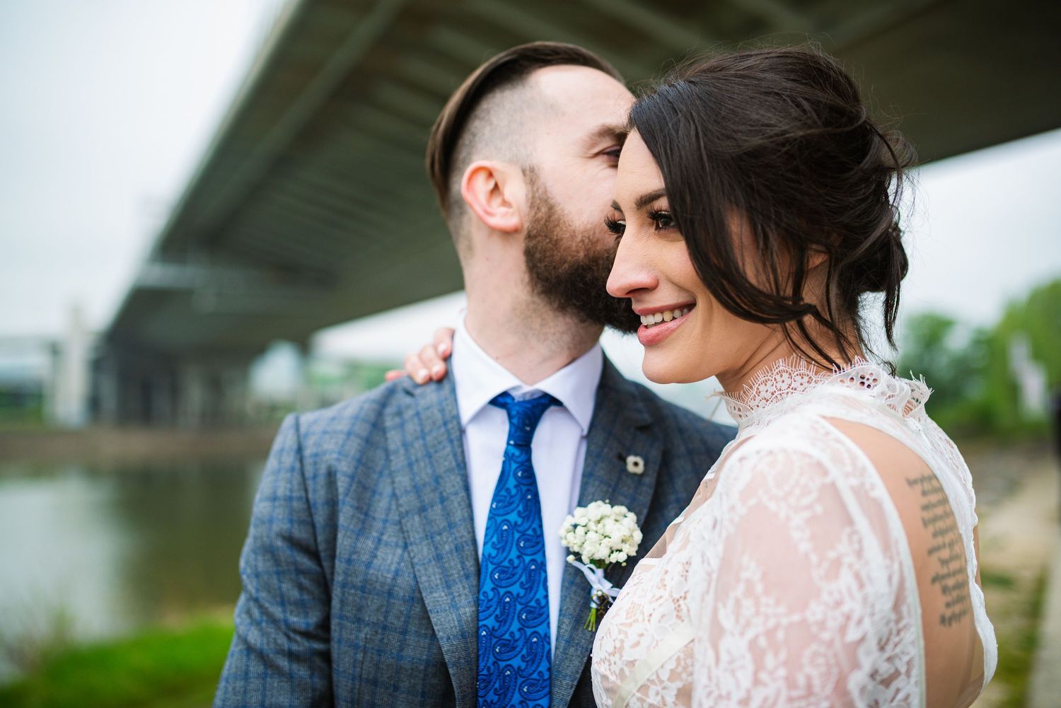 Bearded groom tenderly kissing bride near river with bridge in background during outdoor wedding photos.