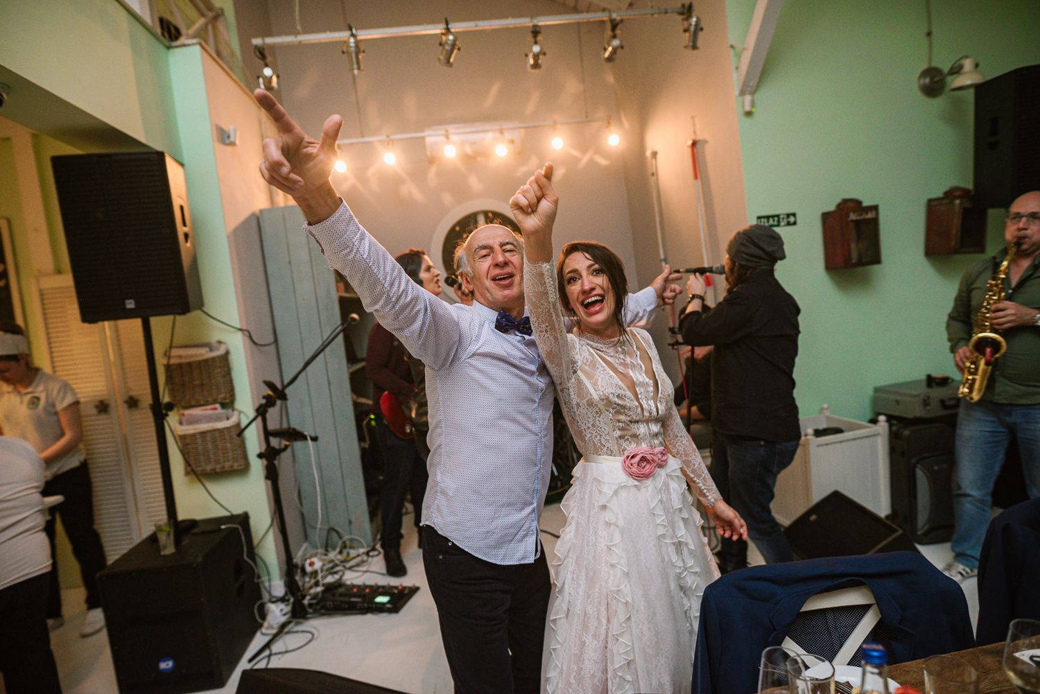 Joyful couple dances with arms raised at wedding reception with live saxophone band.