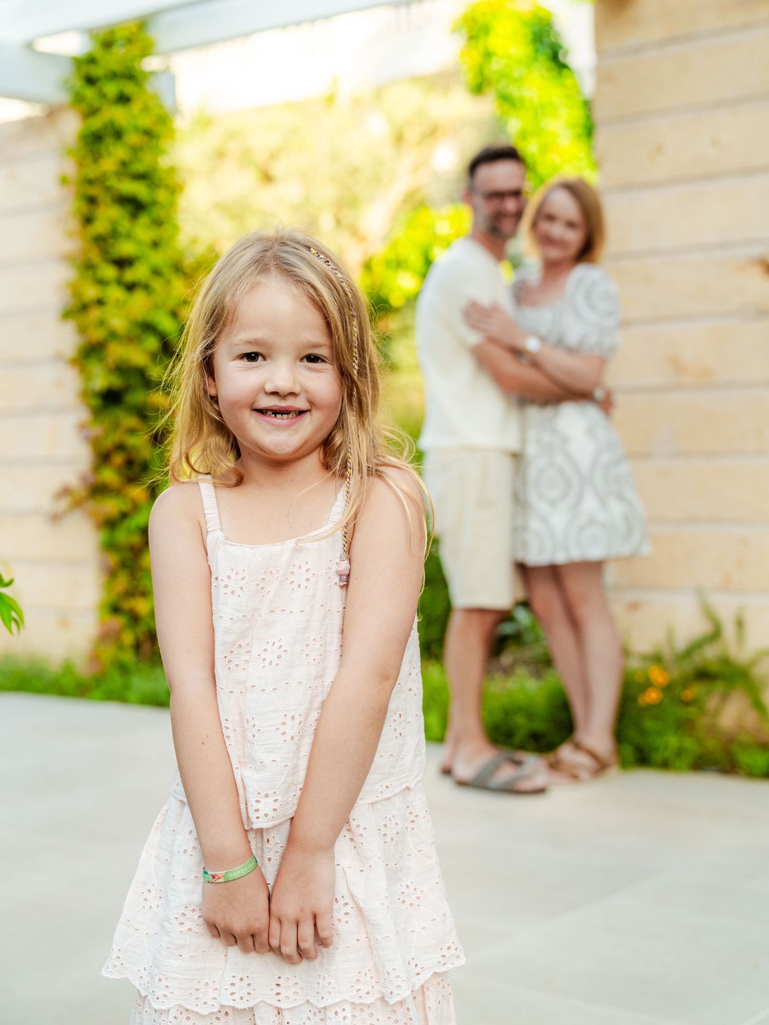Smiling blonde girl in pink dress poses in foreground while parents embrace softly behind her.
