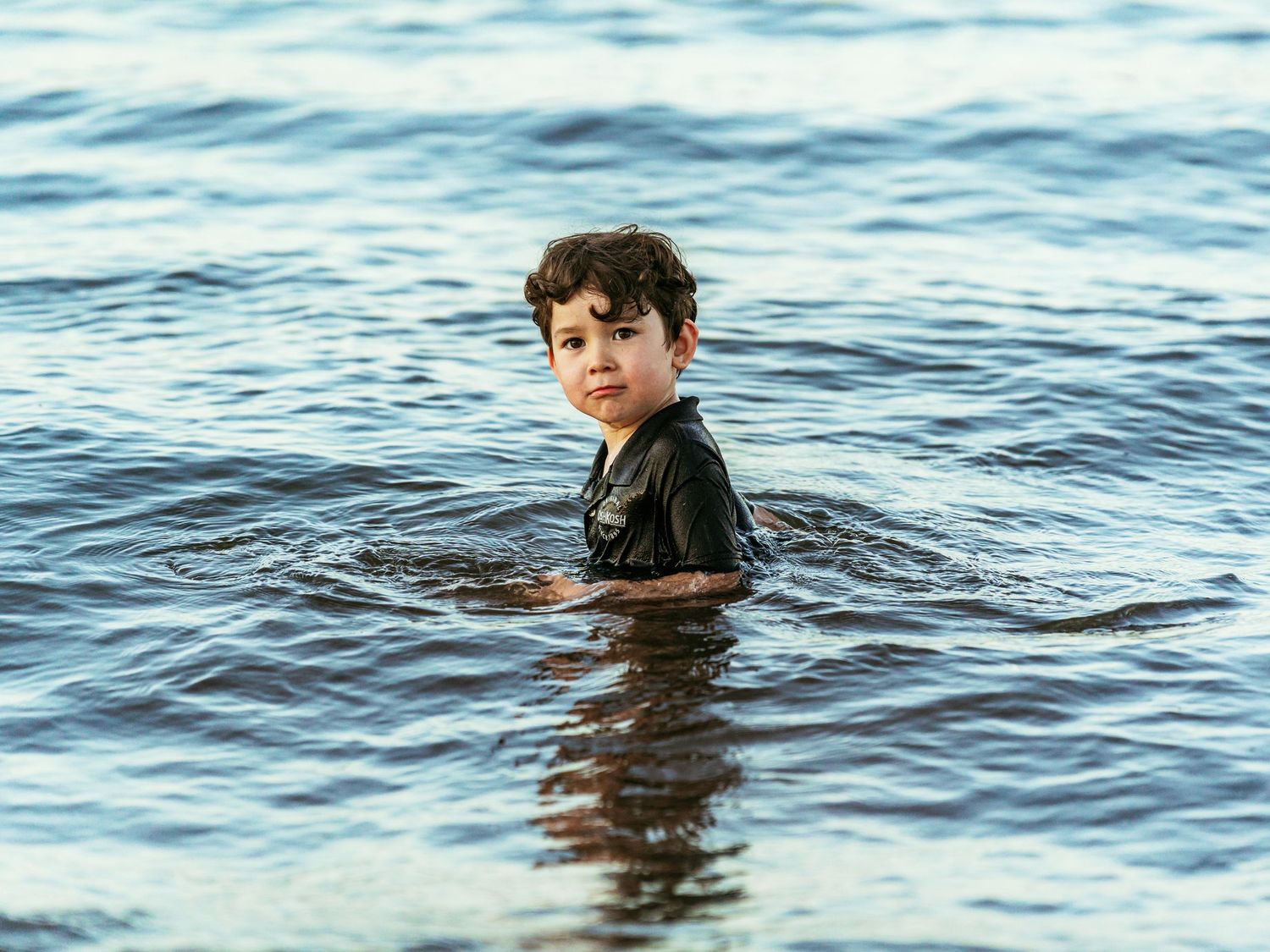Young boy wading chest-deep in calm ocean water looking back at camera.