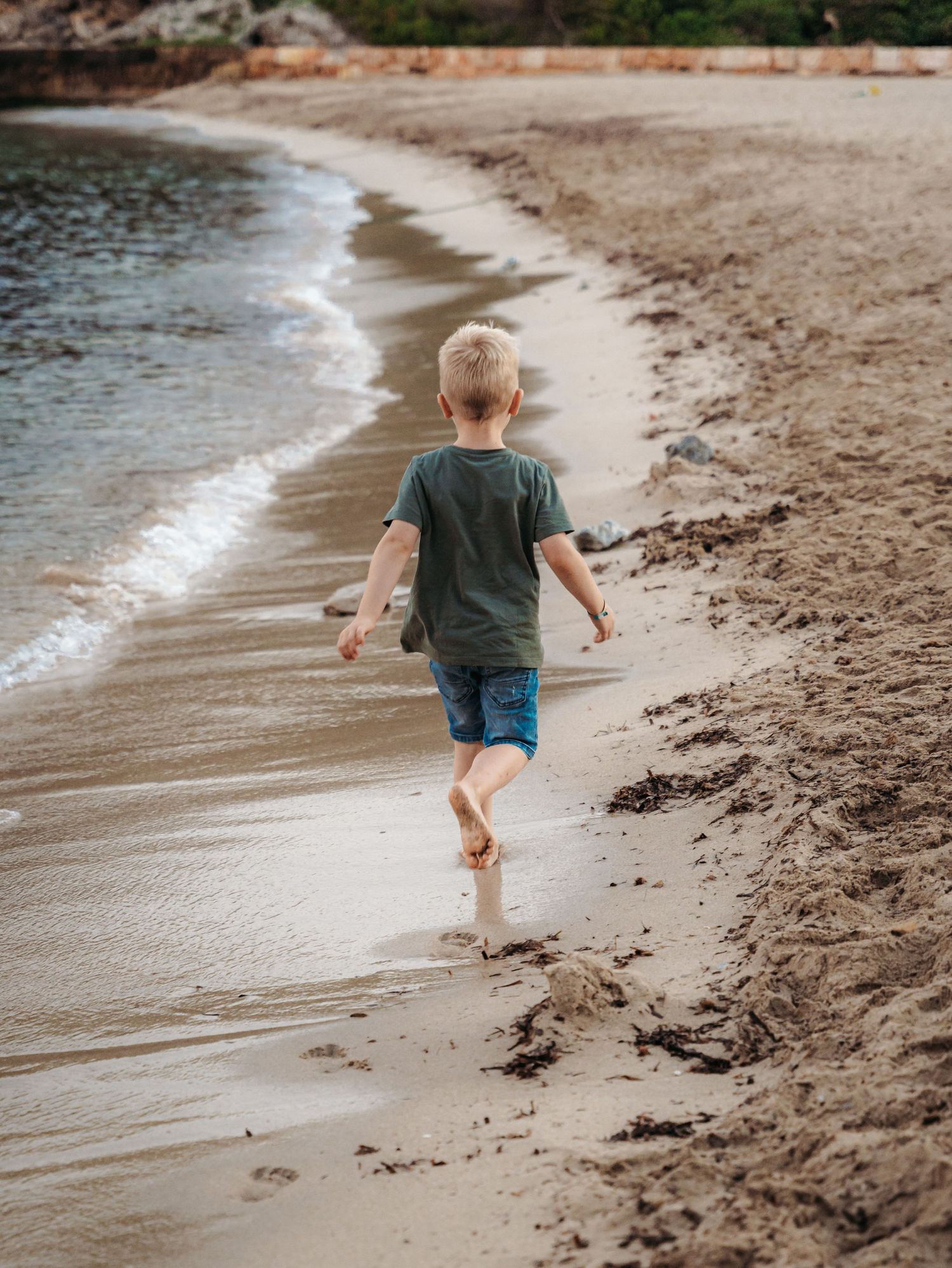Young blonde boy in green shirt running barefoot along sandy beach near ocean waves.