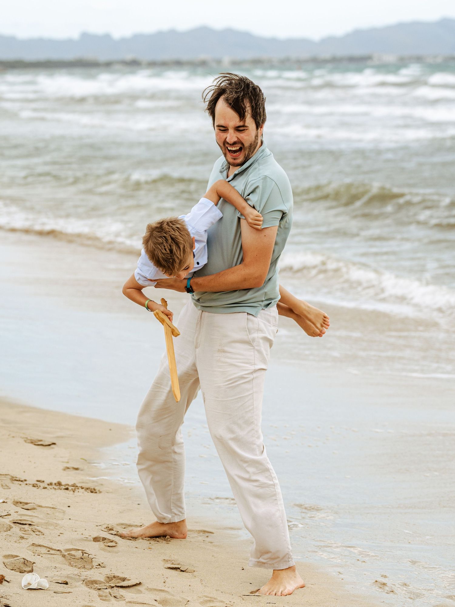 Laughing father carrying young boy with wooden sword on a windy beach near ocean waves.