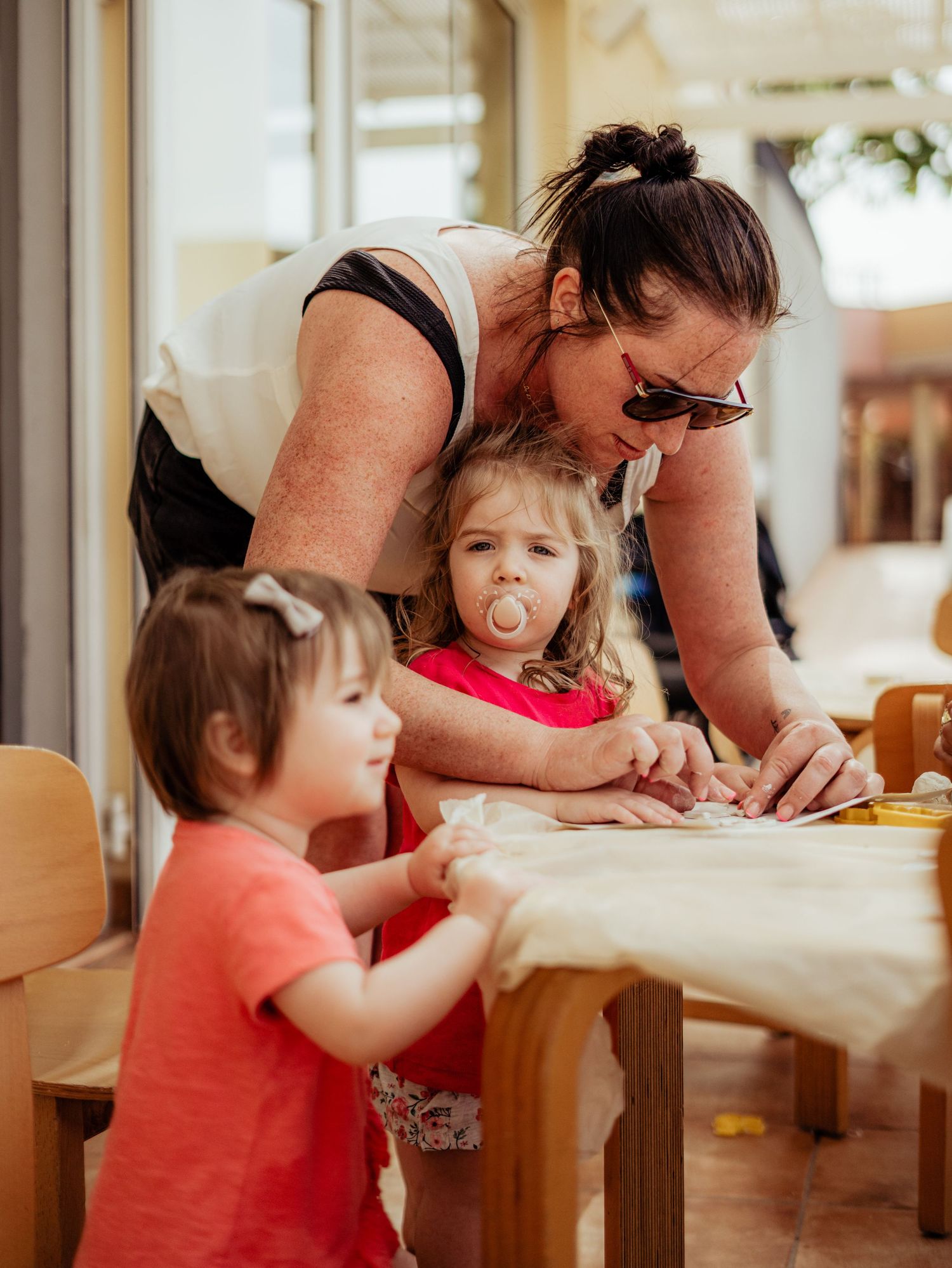 A mother leans over two young toddler girls doing an activity at a wooden table outdoors.