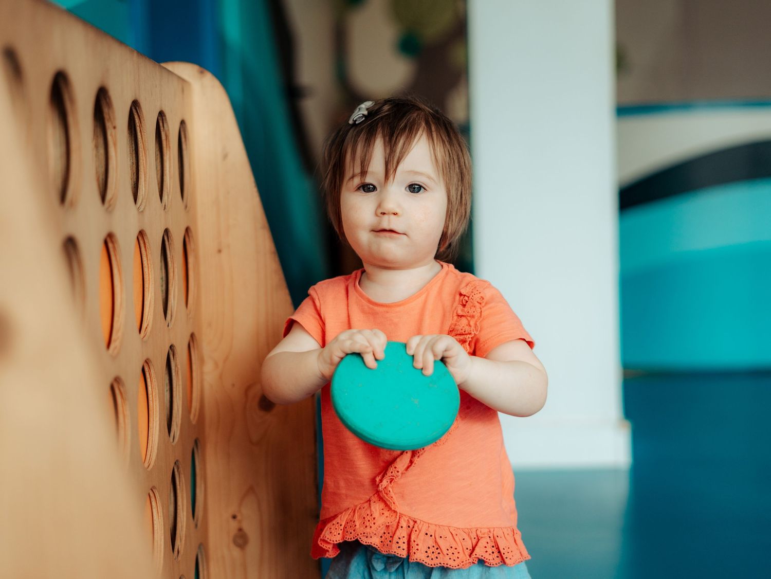 Toddler girl in coral top holding a teal toy disc at a wooden play structure.