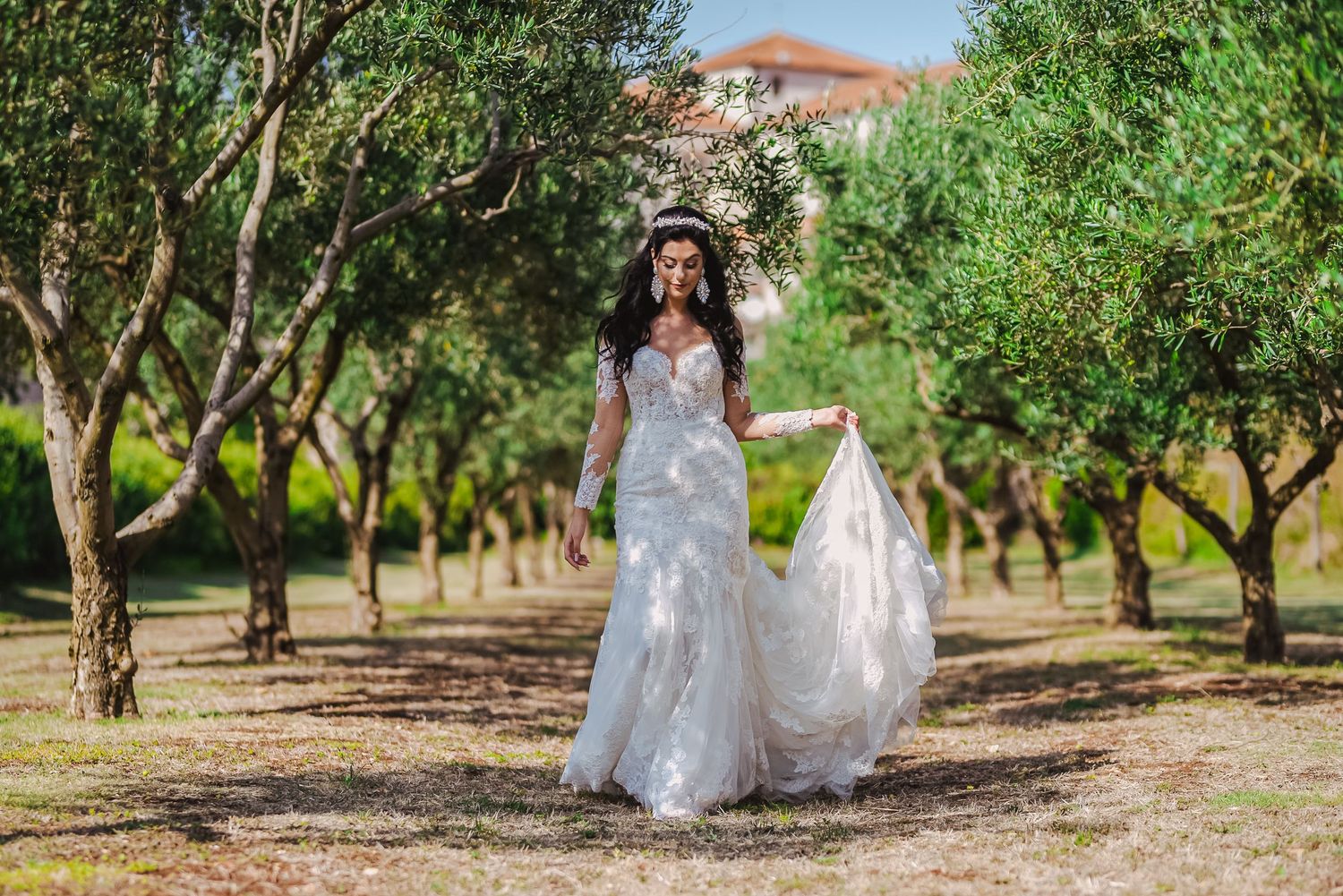 Bride in a long-sleeve lace wedding gown walks through a tree-lined path wearing a floral crown.
