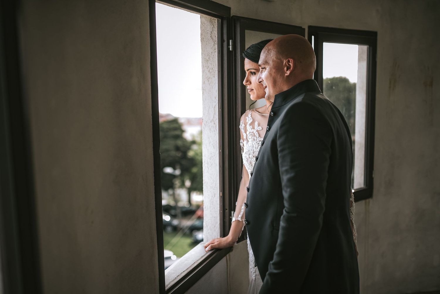Bride and groom standing together gazing out a window in a romantic indoor portrait.